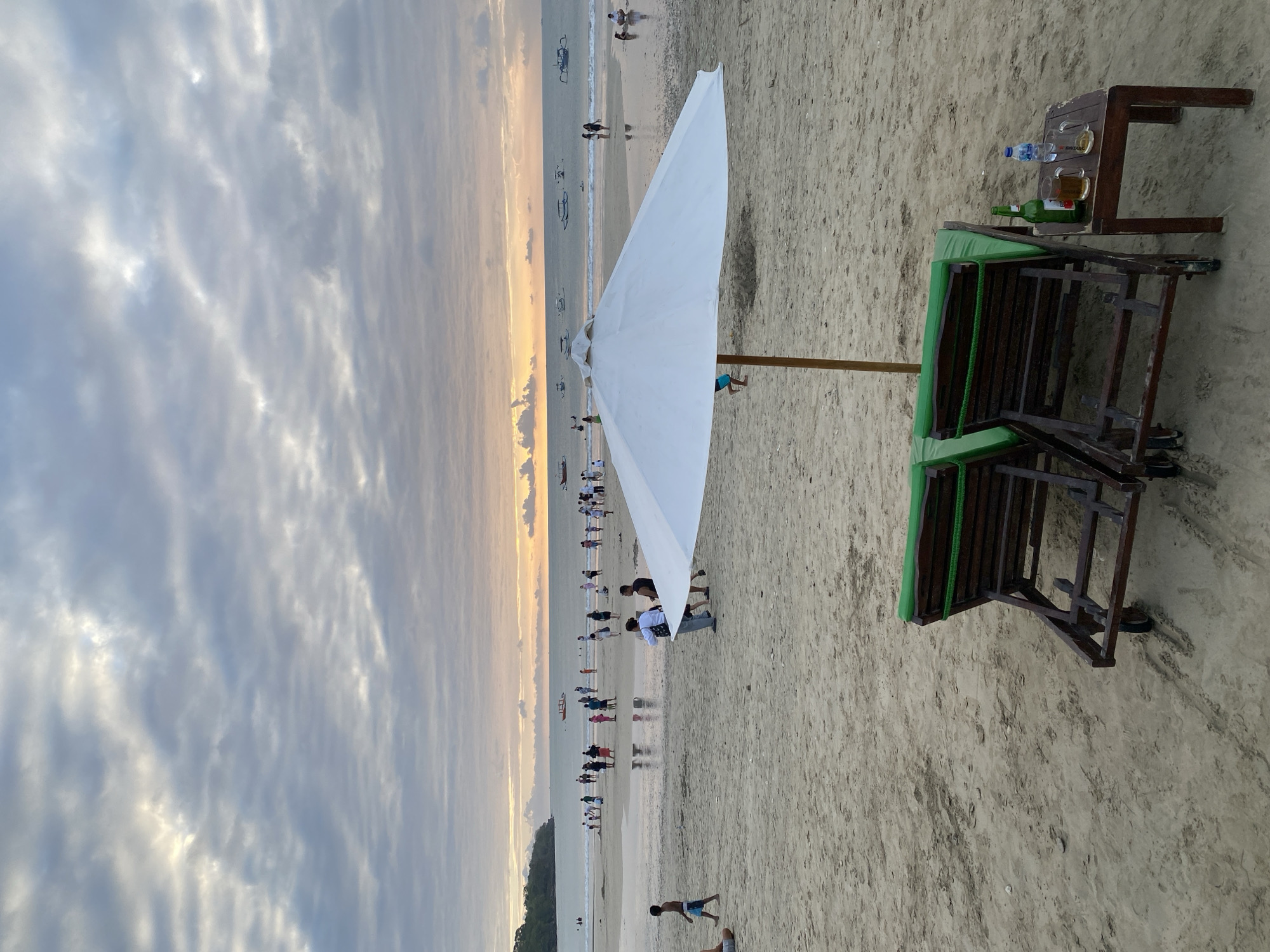 The image depicts a beach scene in Kuta Selatan, Indonesia, during a cloudy sunset. In the foreground, two empty green lounge chairs with wooden frames sit on the sand, accompanied by a small wooden table holding a green beer bottle, two beer mugs, and a bottle of water. A large white umbrella is positioned behind the chairs. The middle ground shows a wide stretch of sandy beach with numerous people scattered across it, some walking, some standing in shallow water, and some gathered in groups. Several boats are visible on the calm ocean in the background, with a hazy orange glow from the setting sun visible on the horizon. The sky is filled with a mix of gray and white clouds, with some patches of brighter sky where the sun is setting. A small, tree-covered landmass is visible on the far left of the image. The overall atmosphere is that of a popular tourist destination winding down for the day.