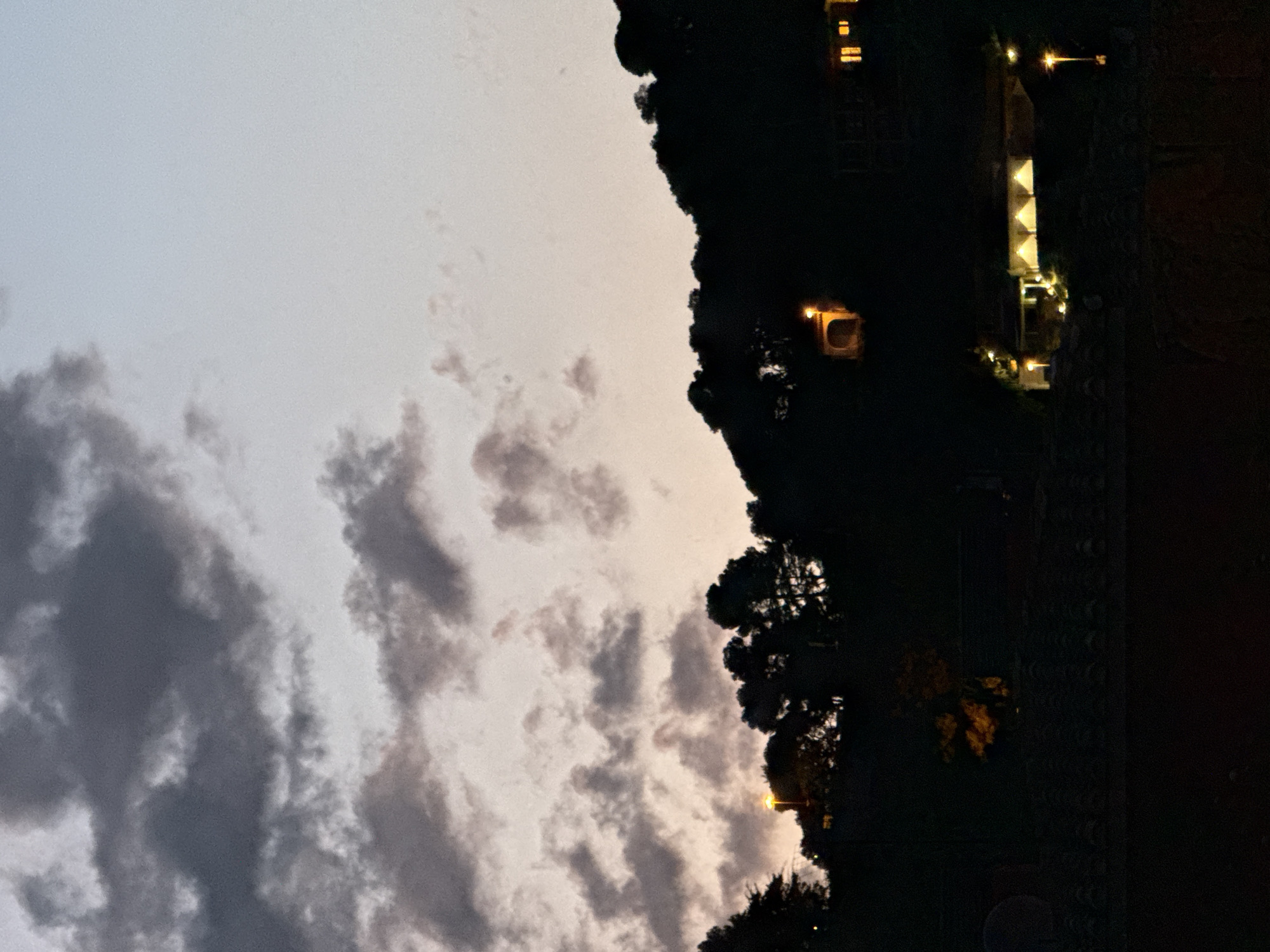 This is a photograph of a hillside at dusk in Blanes, Spain. The sky is a pale gradient of blue and pink, with dark clouds gathered in the upper left quadrant. Silhouetted against the sky are the dark shapes of trees and the contour of the hillside. Several structures are visible on the hillside, illuminated by artificial lights. In the middle ground, a building with a series of illuminated arches is visible, along with several streetlights. Higher up on the hill, another building with illuminated windows can be seen. A single streetlamp emits a bright light in the lower right portion of the frame. The foreground is dark and indistinct, appearing to be a tiled roof or ledge.