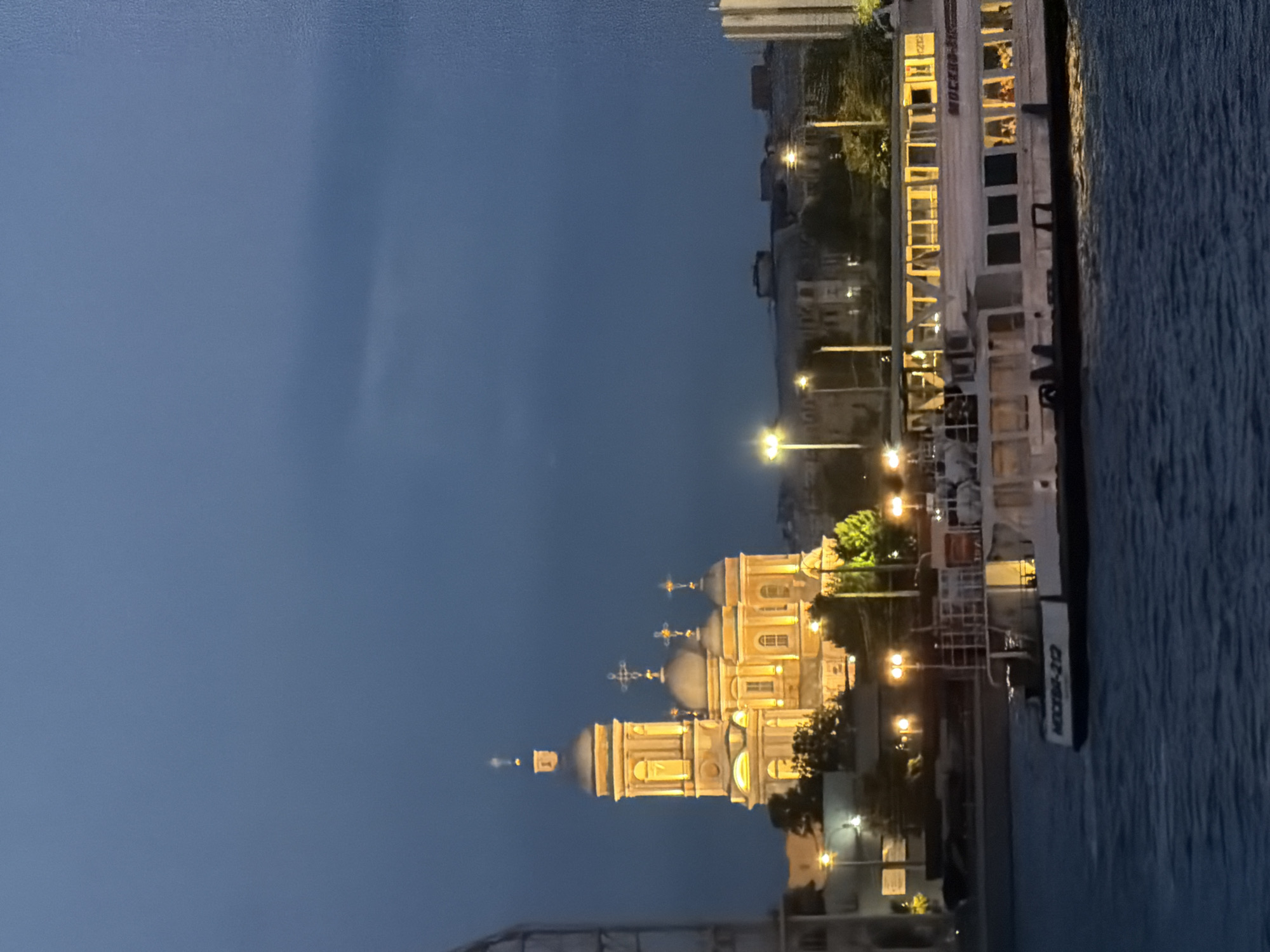 The image captures a twilight scene of Saint Petersburg, Russia, with a prominent illuminated church dominating the left side of the frame. The church features multiple domes and intricate architectural details, all bathed in a warm, artificial light against the darkening blue sky. In the foreground, a white passenger boat with yellow-lit windows glides across the water. The boat has text on its side that appears to read "МОСКВА-212". The water is dark and somewhat choppy, reflecting the lights from the city. Along the waterfront, there are visible streetlights and the silhouettes of other buildings, suggesting a developed urban environment. The overall atmosphere is tranquil, with the man-made lights contrasting with the natural fading light of the day.