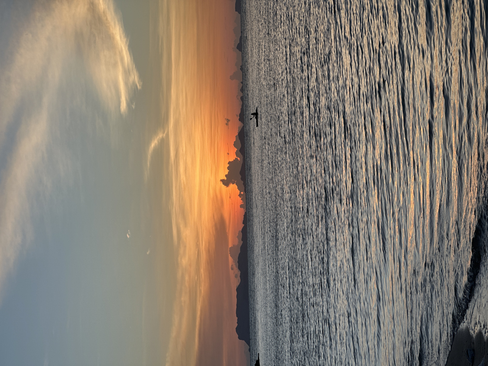 A wide shot captures a serene ocean scene during sunset. The sky is painted with vibrant hues of orange, yellow, and blue, with wispy clouds streaking across. In the distance, a chain of islands forms a dark silhouette against the colorful sky. The calm sea reflects the sky's colors, with gentle waves lapping at the sandy shore in the foreground. A lone kayaker paddles across the water, a small, dark figure against the vast expanse. The image appears to have been taken on the coast of Ko Samui District, Thailand, judging by the geographical features and typical sunset colors.