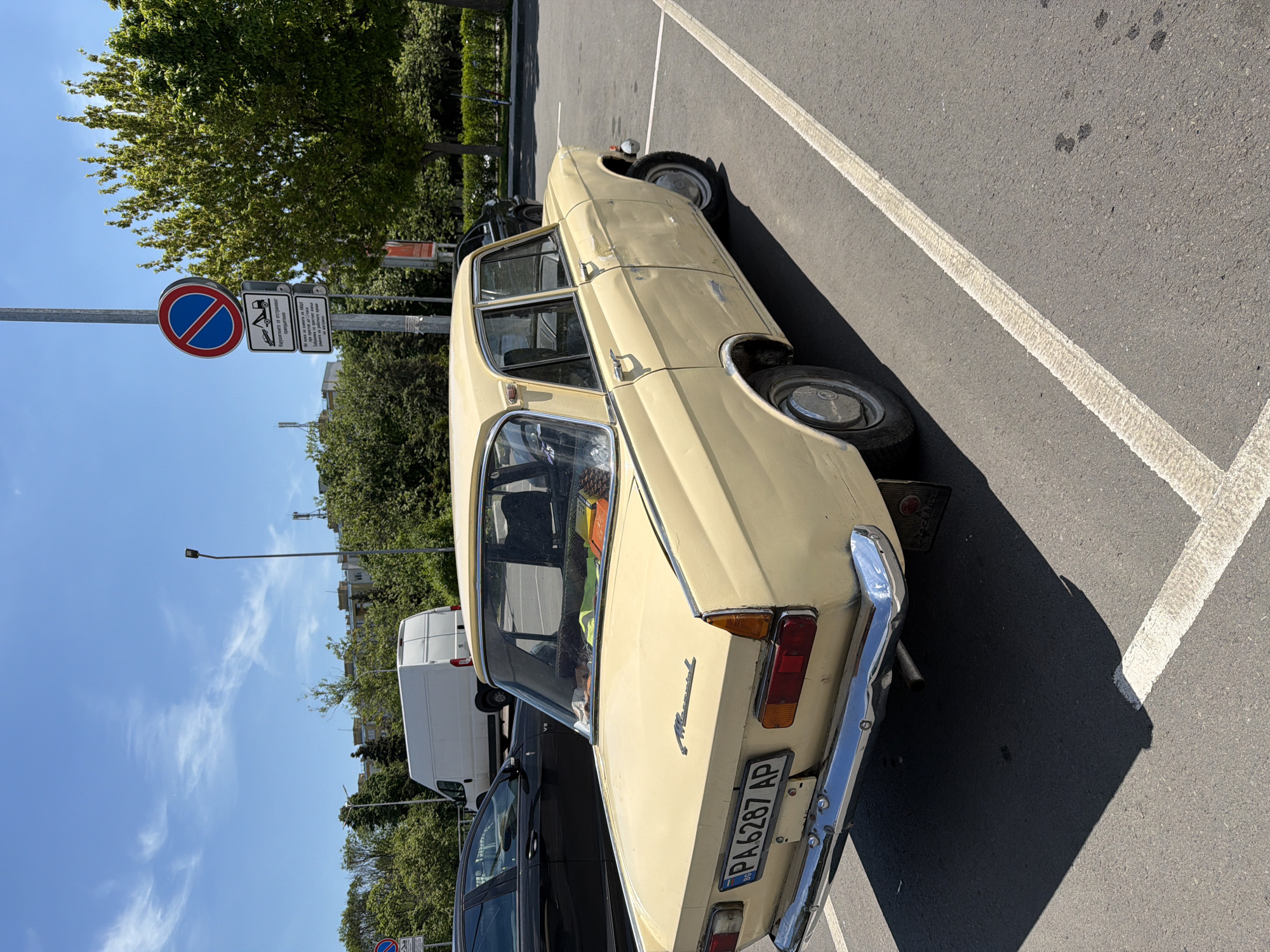 The image shows a beige Moskvich car parked in a parking spot next to a "no parking" sign. The car's license plate reads "PA 6287 AP." The location appears to be in Burgas, Bulgaria, given the license plate format and the Cyrillic writing on the sign. A white van and a black car are parked nearby. The sky is clear, suggesting a sunny day. The scene is a typical urban parking situation.
