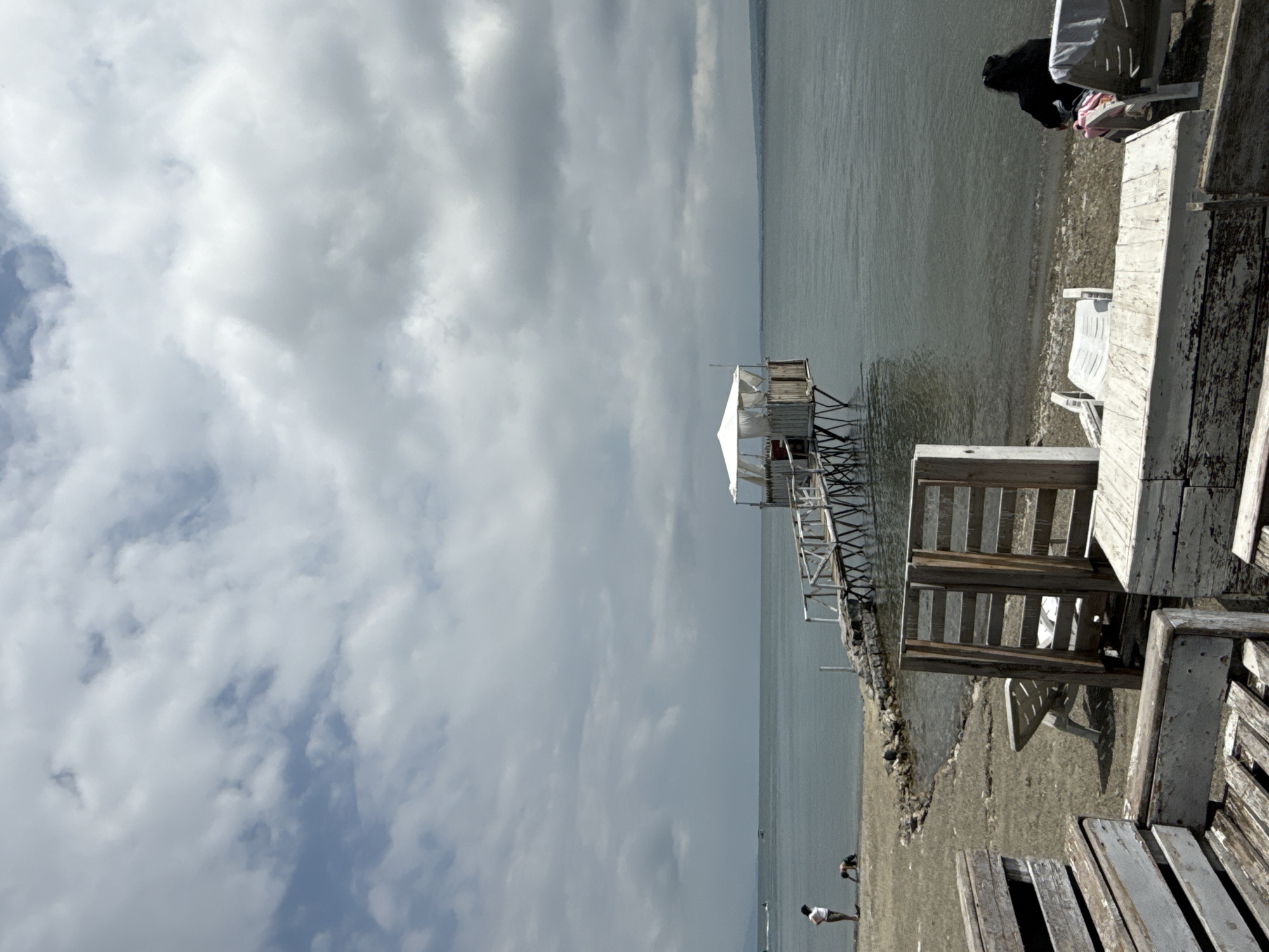 The image depicts a view of the Sevan Lake in Sevan, Armenia. In the foreground, weathered wooden benches and tables are arranged on a gravelly beach. Further back, a wooden pier extends into the water, leading to a small, elevated structure with a white canopy. Two people are visible on the shore near the pier: one standing and looking out at the water, and another person kneeling. The lake stretches to the horizon under a cloudy sky. In the middle ground, several empty lounge chairs are placed on the beach. In the background, a distant shoreline is visible. A person is seated on a lounge chair on the right side of the image, facing the water.