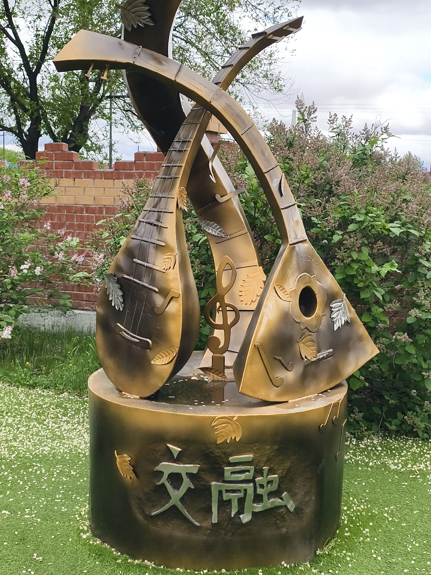 The image shows a sculpture in Blagoveshchensk, Russia, depicting two stylized stringed instruments resembling balalaikas intertwined. The sculpture is made of metal, with a gold and bronze finish. The instruments are adorned with metal leaves and musical notes. A metal treble clef is positioned between the instruments. The sculpture rests on a cylindrical base with Chinese characters on its surface. The sculpture is located outdoors on a green lawn scattered with small white flowers. In the background, there is a brick wall, a tree, and bushes. The sky is overcast.