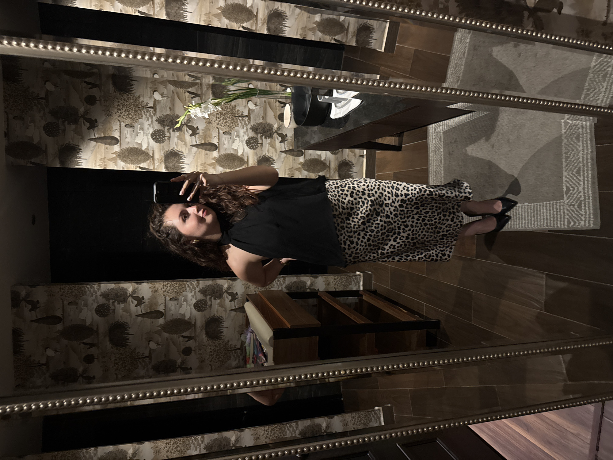 A woman with curly hair, wearing a black top and leopard print skirt, is taking a selfie in a mirror. The mirror's frame is decorated with small beads. The reflection shows a room with wallpaper featuring a bird and tree pattern. A vase with white flowers sits on a table. A dark rug is on the wooden floor. A wooden structure with a cushion is also visible. The image suggests an interior setting, possibly in a home or boutique with decorative elements. The location is not evident from the image.