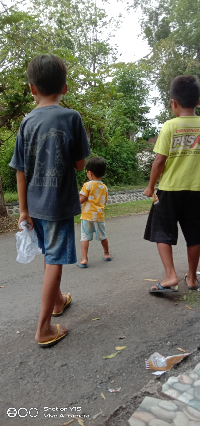 Three boys walk away from the camera on a narrow road. The boy in the foreground wears a blue t-shirt and denim shorts, carrying a plastic bag. The middle boy wears a yellow patterned shirt and patterned shorts. The third boy wears a yellow t-shirt and black shorts. All three wear sandals. Trees and greenery line the road. Litter is visible on the ground in the foreground. The image has a watermark for a Vivo Y15 camera.