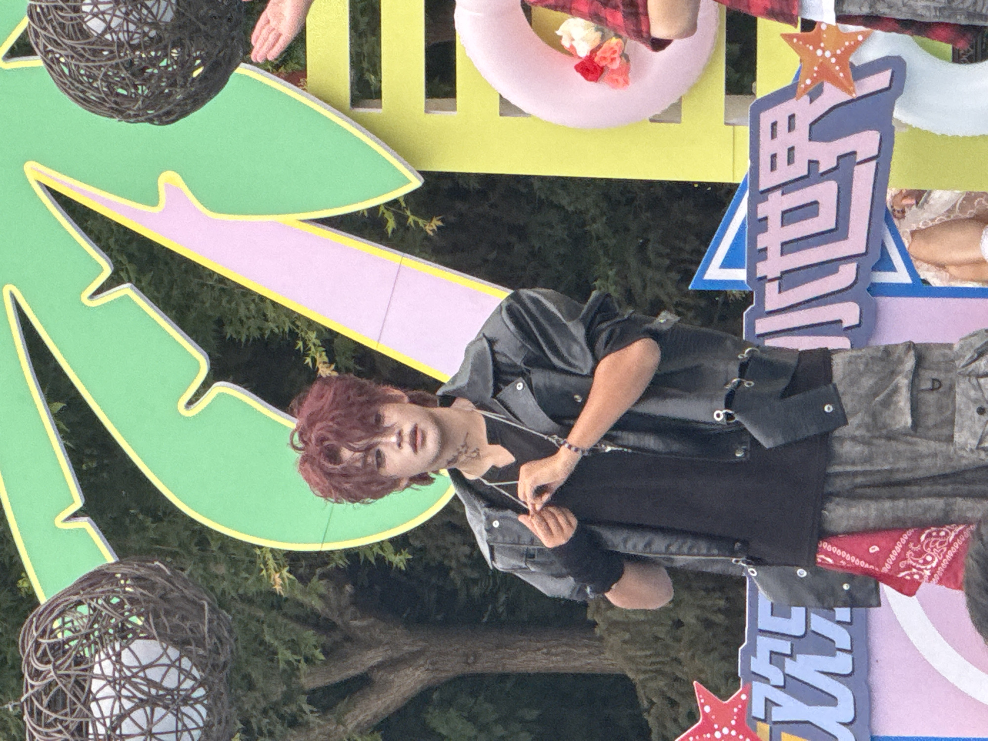 In a city scene in Taihe District, China, a young person with reddish-brown hair and makeup stands, wearing a black t-shirt, a black leather jacket, and camouflage pants. They are holding the string of a necklace and have a beaded bracelet on their wrist. A red bandana is tied to their belt loop. In the background, there is a large, green, palm-tree-shaped decoration with pink and yellow accents. To the left, a woven sphere hangs from a wire. To the right, a pink inflatable ring with flowers is visible, and a person in a red plaid shirt is partially seen. The scene appears to be outdoors, with trees and foliage in the background. Two signs with Chinese characters are present in the foreground.