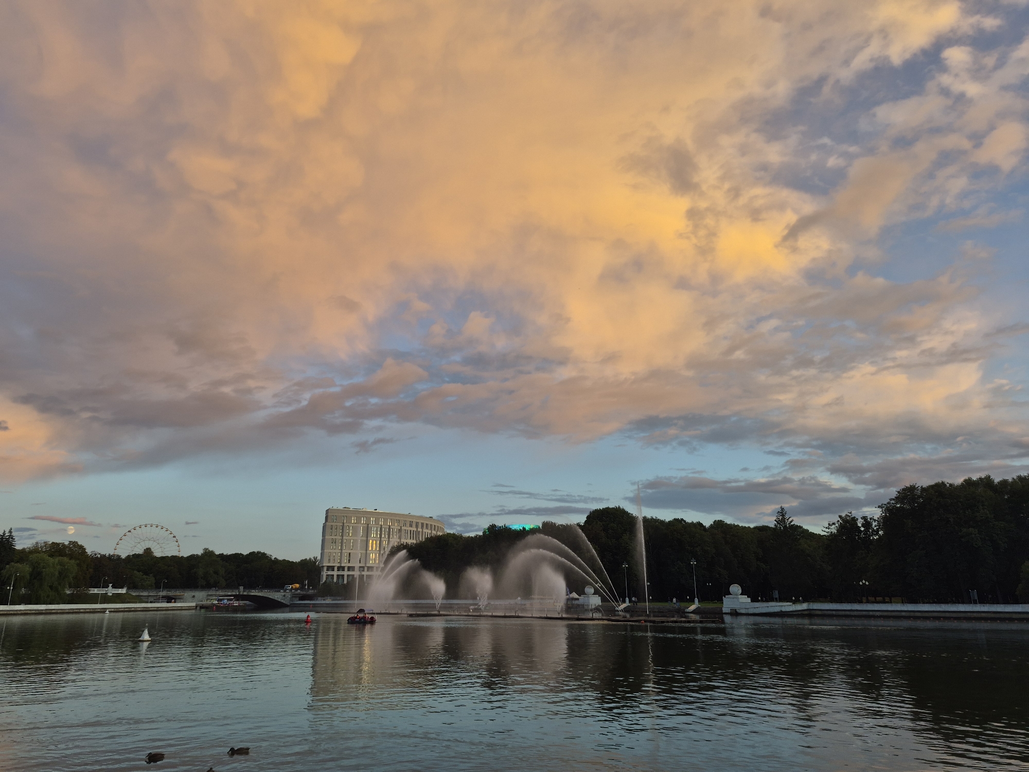 A wide-angle photograph captures a lake in the city of Minsk, Belarus, at dusk. The sky above is filled with dramatic clouds, tinged with orange and yellow from the setting sun, contrasting with patches of pale blue. On the far side of the lake, a modern, circular white building stands prominently, its windows reflecting the sky. Adjacent to the building, a large, multi-jet fountain sprays water high into the air, creating a dynamic spectacle. A Ferris wheel is visible in the distance, nestled behind a line of dark green trees that form the backdrop. The water of the lake is calm, with ripples reflecting the muted colors of the sky and surrounding scene. Several small boats, including a red pedal boat and a white sailboat, are visible on the water. In the foreground, two ducks swim peacefully. The overall impression is one of serene urban twilight.