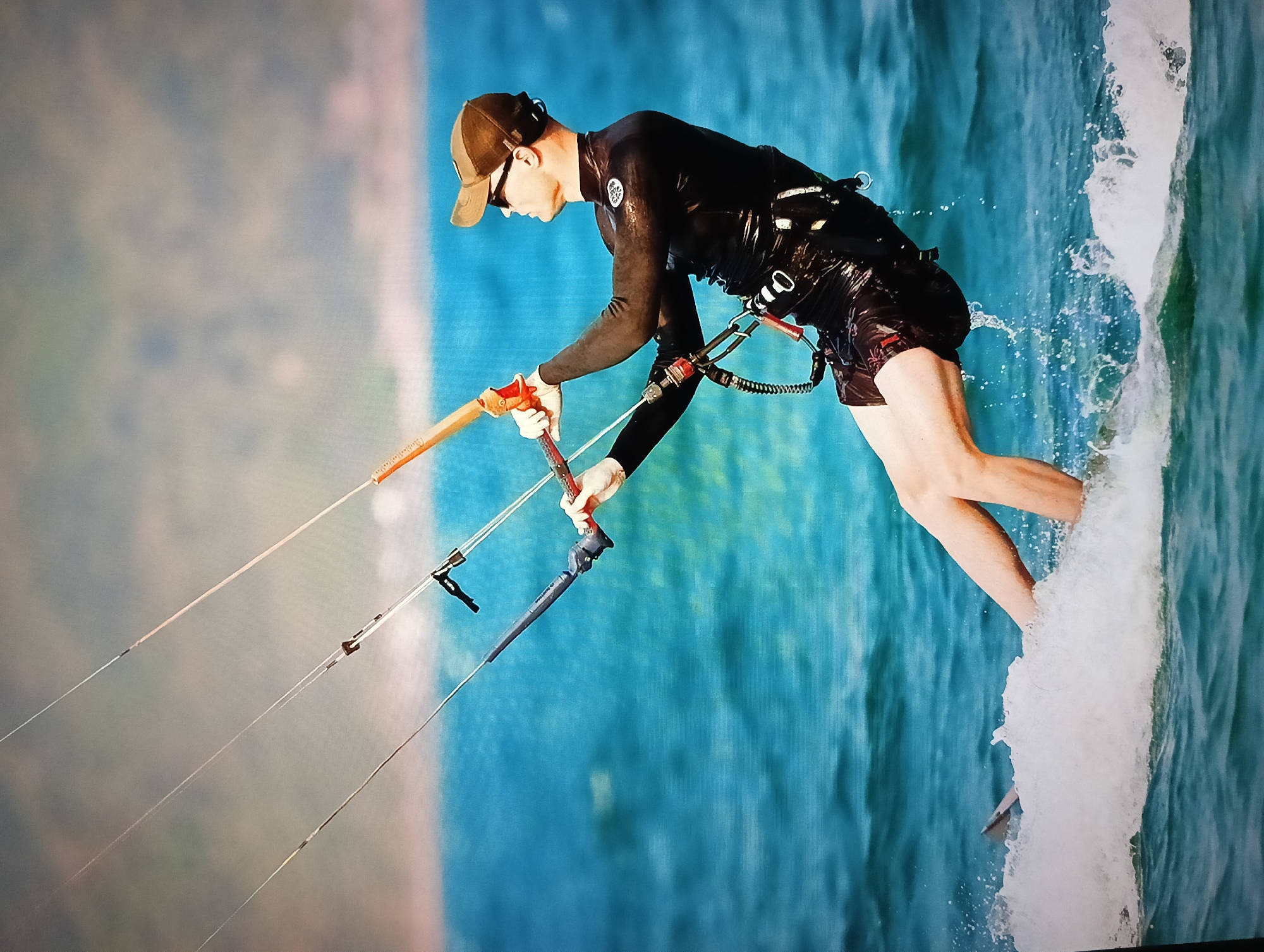 A man is kitesurfing on turquoise water. He is wearing a black long-sleeved wetsuit, black shorts with a design, a brown baseball cap, and sunglasses. He is gripping a control bar attached to kite lines. White water sprays off the side of his board. The location is Nha Trang Ward, Vietnam, discernible by the coastline in the background.