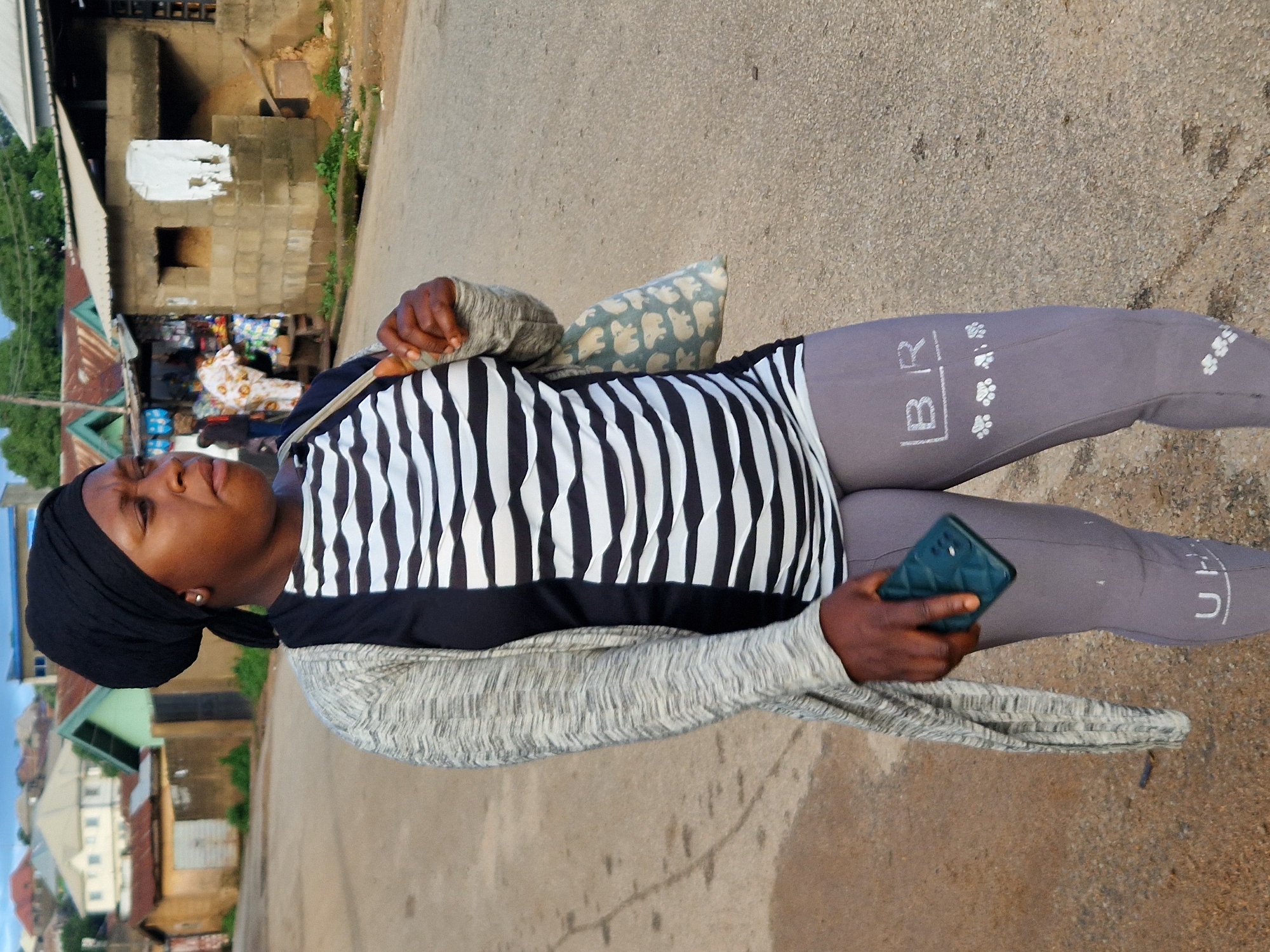 In Kabong, Nigeria, a woman walks on a paved road, holding a dark green smartphone in her right hand. She wears a black headscarf, a black and white horizontally striped long-sleeved shirt, and grey leggings with white text and paw print graphics. A light grey, textured cardigan is draped over her left arm. She carries a light-colored shoulder bag with a pattern of light grey polar bears. In the background, buildings with corrugated metal roofs and concrete structures are visible, along with some vegetation. The sky is overcast.