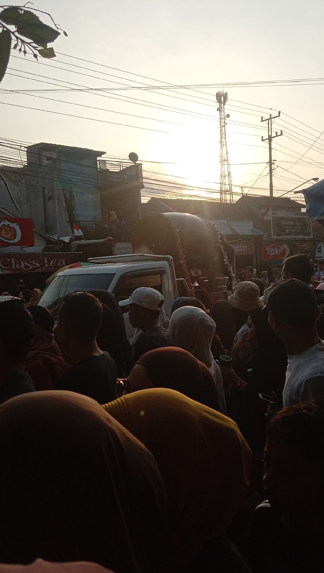 The image depicts a street scene in Jatisawit, Indonesia, at sunset. A large crowd of people are gathered, many of whom are wearing headscarves or hats. In the foreground, several individuals are visible from the back, with their heads bowed. A white pickup truck is parked in the middle ground, and a large cylindrical object adorned with red and white streamers is mounted on a raised platform behind it. This object appears to be a prominent feature of a procession or event. The background shows buildings, power lines, and a telecommunications tower silhouetted against the bright sky. The lighting suggests a warm, late afternoon or early evening atmosphere. The overall impression is of a community gathering or celebration.