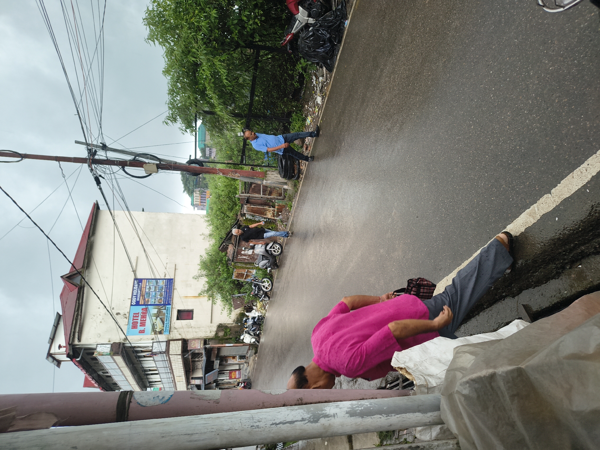 The image depicts a street scene in Dharamshala, India, on a cloudy day, with the pavement appearing wet. In the foreground, a person wearing a bright pink shirt and grey pants is leaning against a pole. To the right, a man in a blue shirt and dark jeans walks down the street. Further down the street, another man in a black t-shirt and blue jeans is also walking. Several motorcycles are parked along the side of the road, and some are covered in black plastic bags. A multi-story building with a red roof is visible in the background, with a sign advertising "HOTEL B. MEHRA" and "OGGY HOLIDAYS DHARAMSHALA". The overall environment suggests a somewhat cluttered urban setting with some commercial establishments.