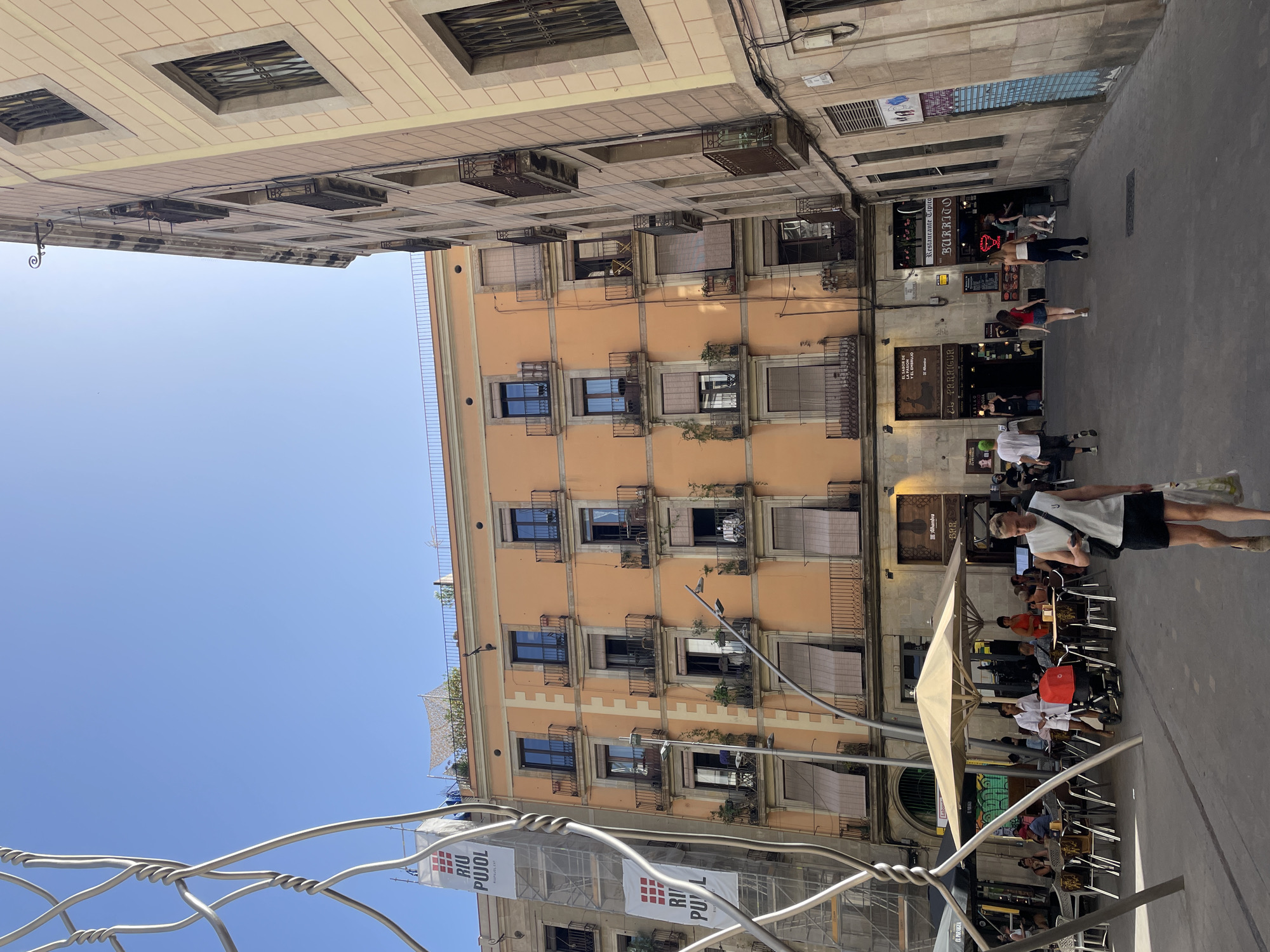 This is a vertical, eye-level shot of a street scene in Barcelona, Spain. The image is dominated by the warm, peach-colored facade of an apartment building on the right, with multiple floors of windows and balconies visible. Below, a street-level view reveals several businesses, including "Restaurante Tipico BURRITO" and "EL PARAUGEA" with signage like "Alhambra BAR." People are seated at tables outside some of these establishments, enjoying the day. A large umbrella shades a seating area. The sky is a clear, bright blue, suggesting a sunny day, likely mid-morning or early afternoon due to the sharp shadows.
On the left side of the frame, another building's facade is visible, constructed with lighter-colored stone or plaster and featuring windows with metal grates. In the foreground, a modern sculpture made of twisted metal wire creates a textured foreground element, partially obscuring a banner with red and white text that appears to read "RIU PUJOL." Several people are walking on the cobblestone sidewalk. The overall atmosphere is lively and typical of a European city during daytime hours.