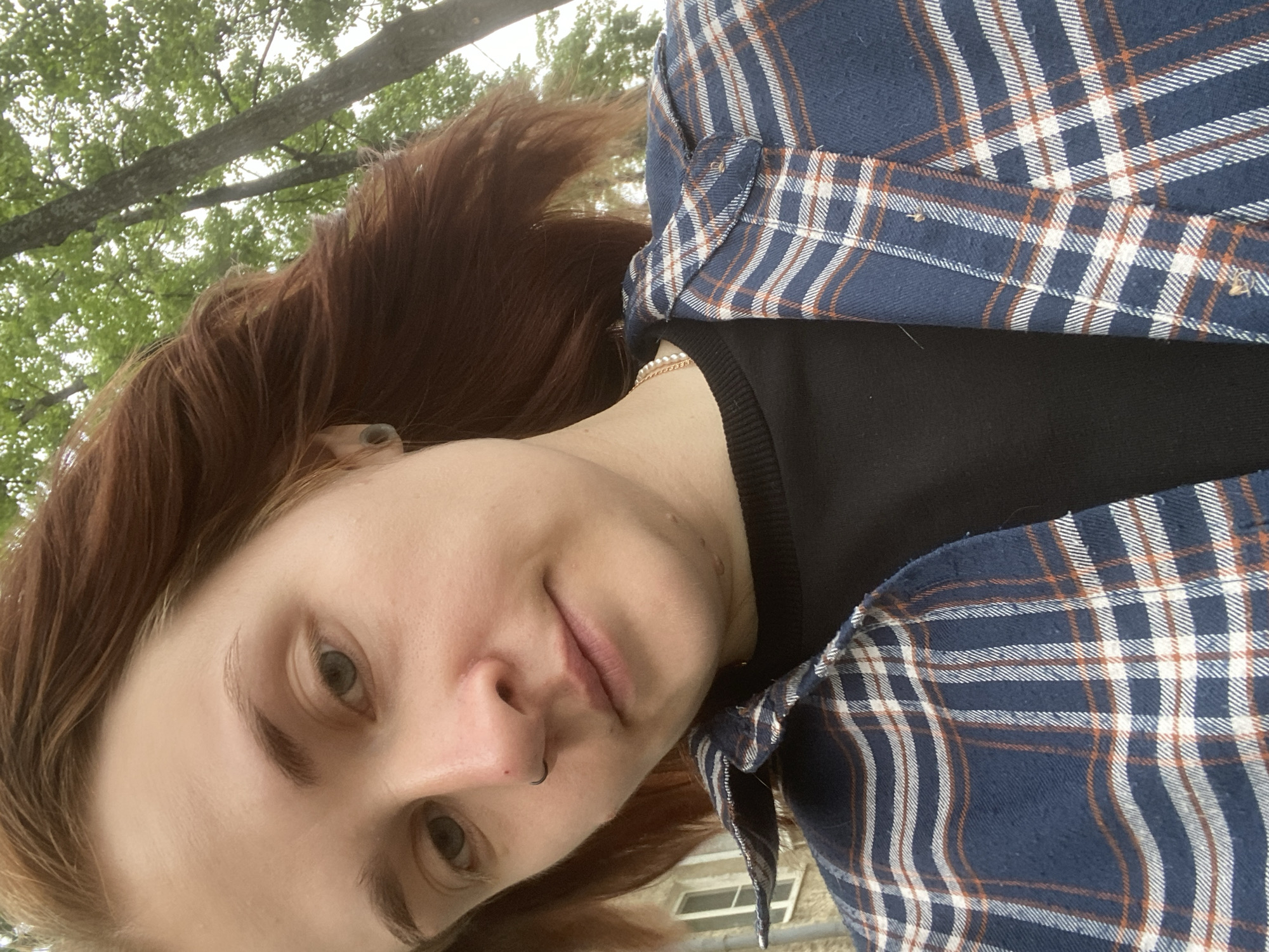 A close-up, selfie-style photograph shows a person with auburn hair against a backdrop of greenery and trees. The individual is wearing a blue and white plaid shirt over a black t-shirt, with a pearl necklace visible. They have a nose ring and an ear gauge. The lighting suggests an outdoor setting, possibly during the daytime. The background includes trees with green leaves and a building with windows. The photograph appears to have been taken in Chaikovsky, Russia.