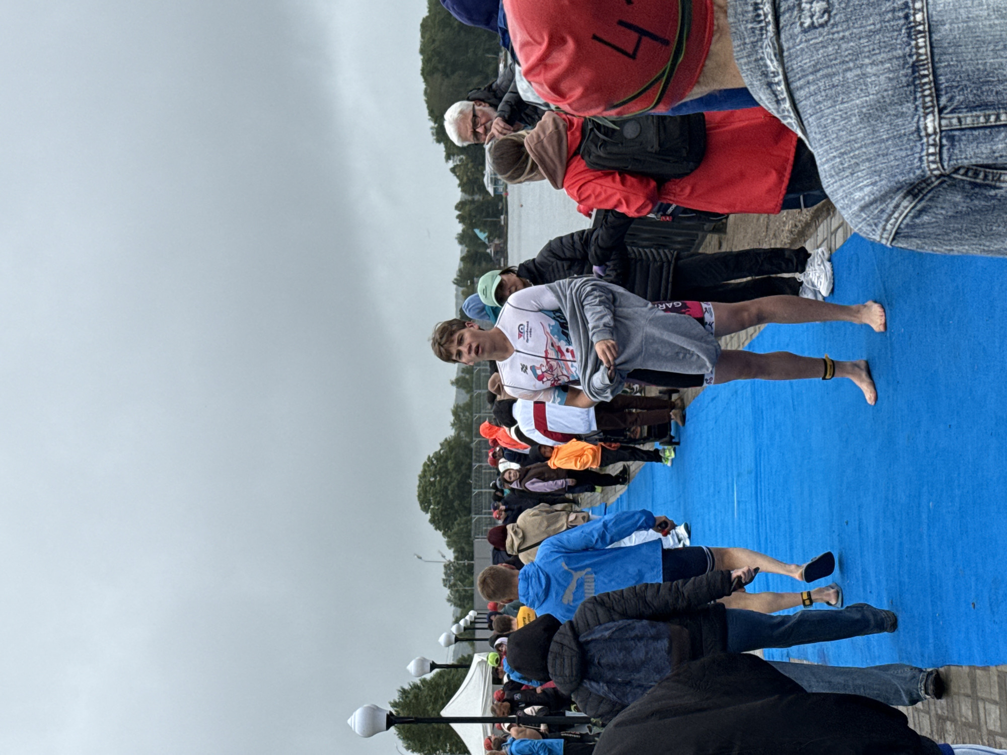 The image depicts an outdoor scene on a cloudy day, likely in Votkinsk, Russia, as suggested by the context. A group of people are walking along a blue carpet laid on a paved surface. In the foreground, a young man wearing a triathlon suit with a gray jacket slung over his shoulders stands facing forward, barefoot, with yellow bands around his ankles. Behind him, other participants in athletic attire are visible. To the left, a white tent is set up, and lampposts line the walkway. The background shows trees and a body of water, possibly Lake Votkinsk. The overall atmosphere appears to be a pre-event or post-event gathering for a sporting competition.