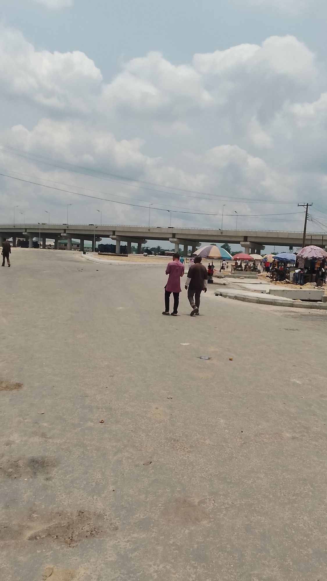 The image captures a daytime scene in Effurun, Nigeria, featuring a wide, unfinished concrete road under a cloudy sky. An elevated highway bridge spans the background. Two men walk away from the viewer on the road, one wearing a magenta shirt, the other in a brown shirt. A smaller figure is visible walking on the far left. To the right, under the bridge, there are small vendor stalls with colorful umbrellas and people gathered around. A power pole is visible near the stalls. The road surface shows patches of discoloration and unevenness.