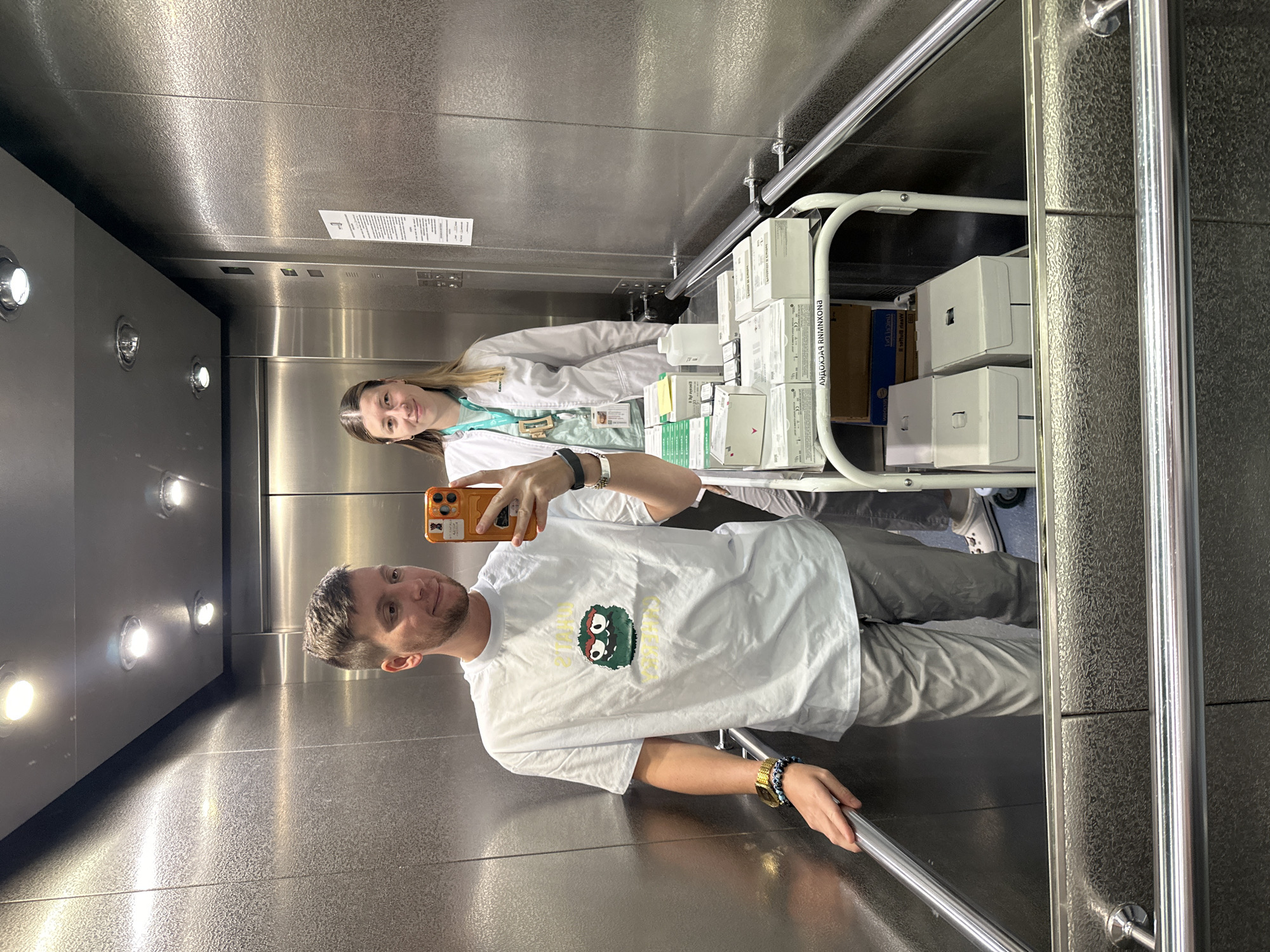 The photo depicts two individuals inside an elevator. A man in a white T-shirt is taking a selfie, while a woman in a lab coat stands behind him, next to a metal cart filled with boxed items. The elevator has a metallic interior. One box has text in Russian, namely, the word "АВОХИНХИМ" written backwards, which suggests that this photo was taken somewhere in Russia. While not apparent, there's a high chance that this image was taken in Moscow due to the high concentration of pharmaceutical and scientific companies in the capital city.