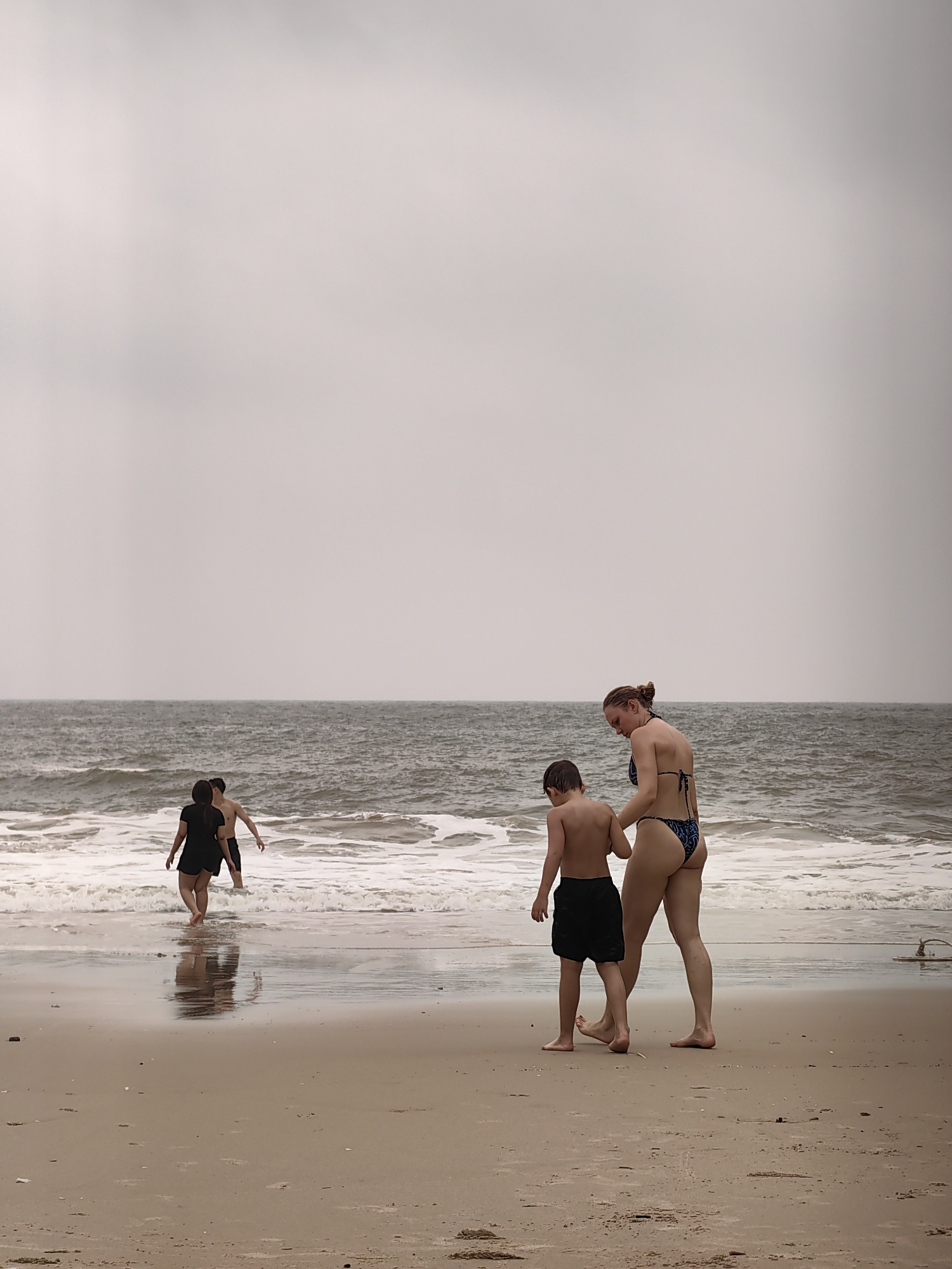 On a beach, a woman in a blue patterned bikini is walking on the beach with a young boy in black shorts, who has short brown hair and a bare torso. They are walking near the water's edge. Two other figures, also partially submerged, are in the water further away. The sky is overcast and gray, and the ocean has white waves. The sand is tan. The scene is on a beach that could be Do Son beach in Hai Phong, Vietnam, although this cannot be confirmed definitively from the image alone.