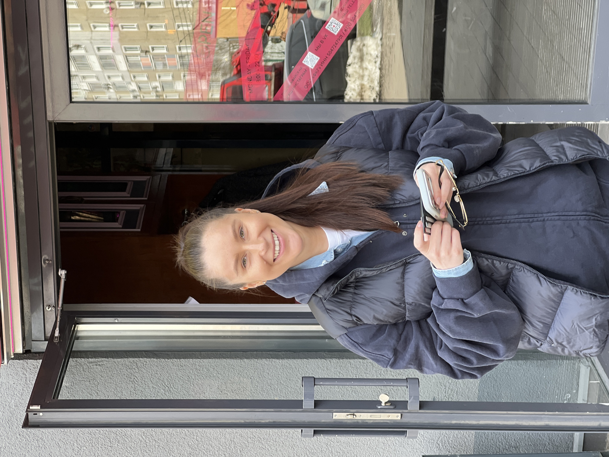 A woman is standing at the entrance of a business establishment in the city of Khimki, Russia. She is wearing a blue quilted jacket, a denim shirt, and a dark-colored sweatshirt. The woman is holding a pair of eyeglasses and a smartphone in her hands. Behind her is a storefront with opening hours displayed on a sign: "MONDAY-FRIDAY 8:00-22:00". A sign with social media information is also visible: "ДПИСЫВАЙСЯ НА НАШ ИНСТАГРАМ" (Subscribe to our Instagram). Legal information is also on the door: "ООО «ФКГ» ИНН 5047247901 / ΚΠΠ 504701001". The storefront has a window reflecting an apartment building across the street. The overall setting appears to be an urban, commercial area.