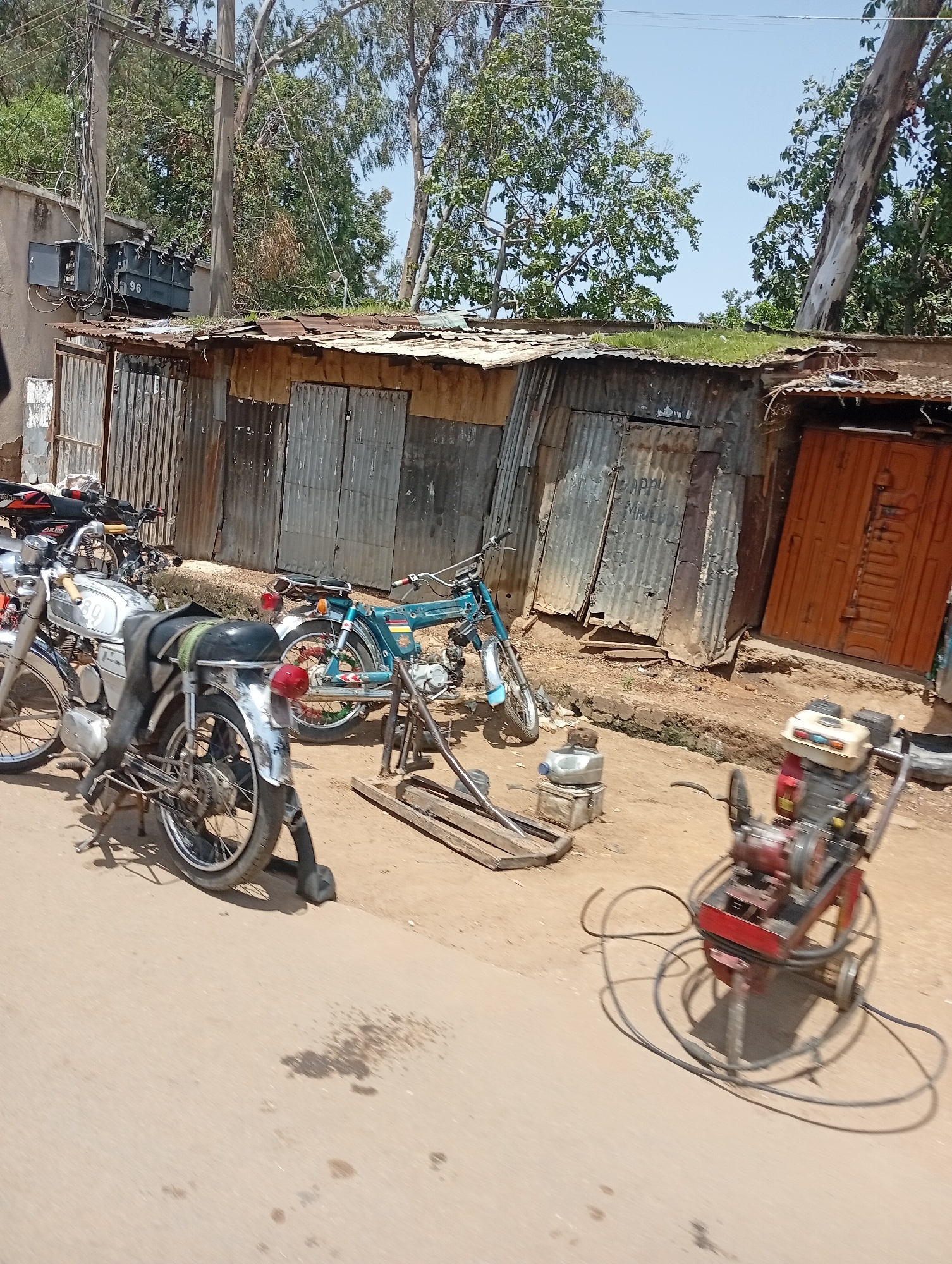 The image captures a street scene in Jos, Nigeria, likely in a commercial or residential area. In the foreground, a light-colored motorcycle is parked on a dirt and paved road surface. A black seat cover is draped over its rear seat. Behind it, a blue motorcycle is also parked. To the right of the motorcycles, a red and black piece of equipment with wheels and various components, possibly a generator or a portable engine, is positioned on the ground.
The background features a long, corrugated metal structure with several doors. One door on the right is painted a reddish-orange, while others appear to be made of unpainted metal. Above the structure, trees with green foliage are visible against a clear blue sky. Electrical poles and wires are also present, suggesting a developed but potentially aging infrastructure. The overall impression is a snapshot of daily life in a functional, perhaps somewhat rustic, urban environment.