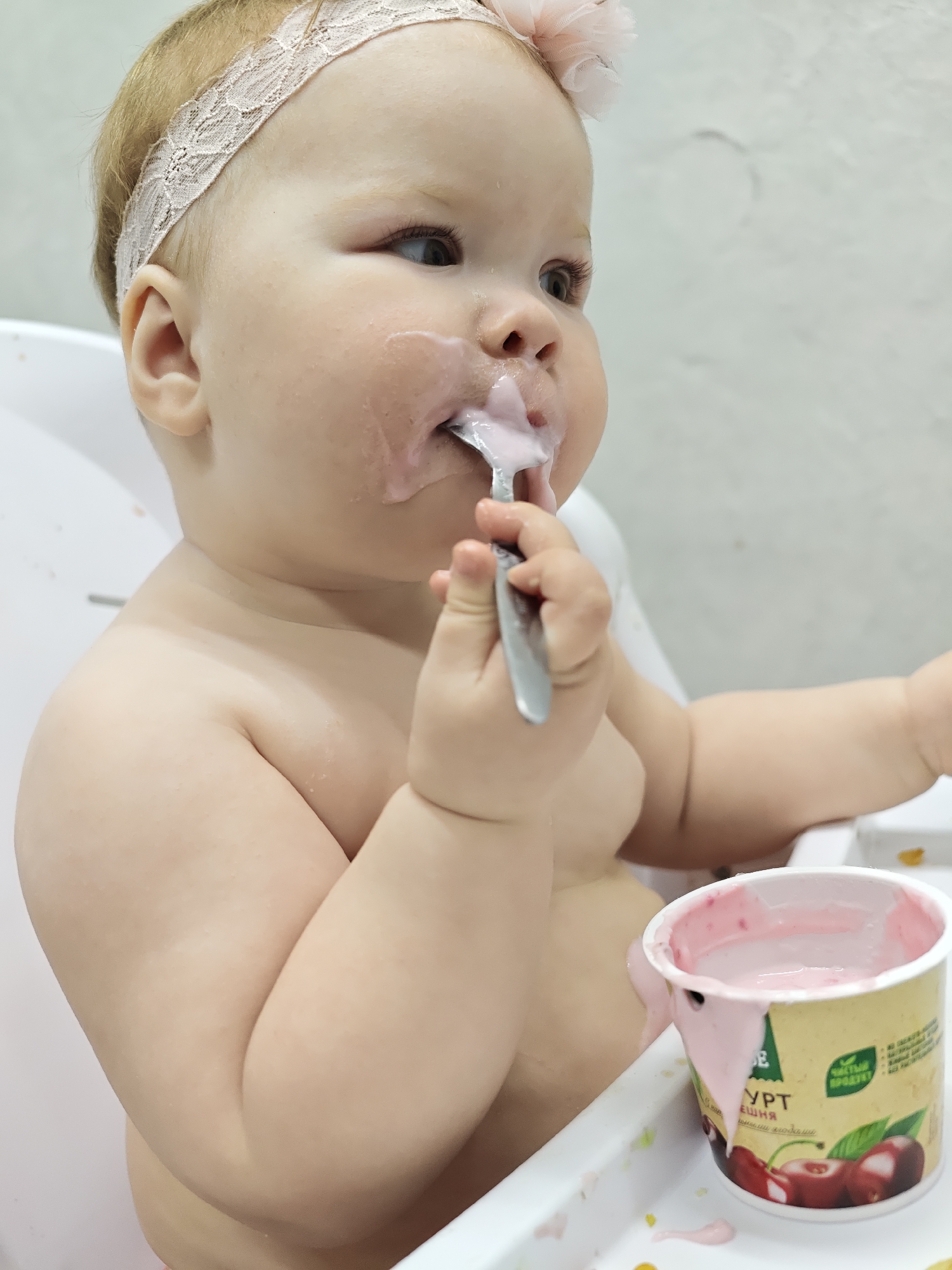 A baby is sitting in a high chair, eating cherry yogurt with a spoon. The baby is fair-skinned with a light pink headband. There is yogurt on the baby's face and the container of yogurt is nearby. The location is likely Chaikovsky, Russia, based on the yogurt container's label in Russian.