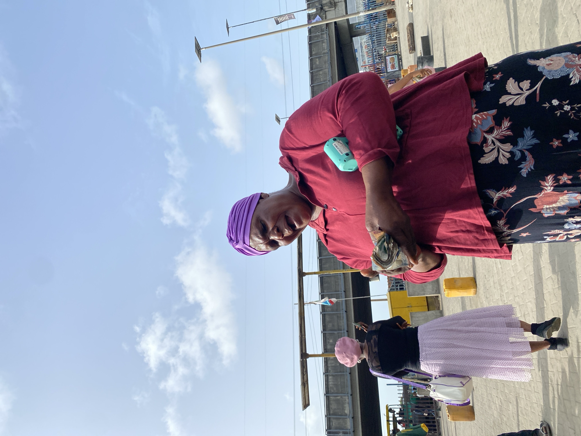 A medium shot of two women in Ado, Nigeria. The woman closest to the camera wears a purple headwrap, a dark red long-sleeved shirt, and a black floral skirt. She holds a wad of Nigerian currency in her hands. Attached to her shirt is a small turquoise device. A second woman stands adjacent, wearing a pink head covering, a black top, and a white polka-dotted skirt. She carries a white purse with a purple strap. The background features a blue sky with scattered clouds, streetlights, metal railings, and stacked containers. The scene appears to be an outdoor marketplace or public area. The foreground shows the concrete ground where they stand.