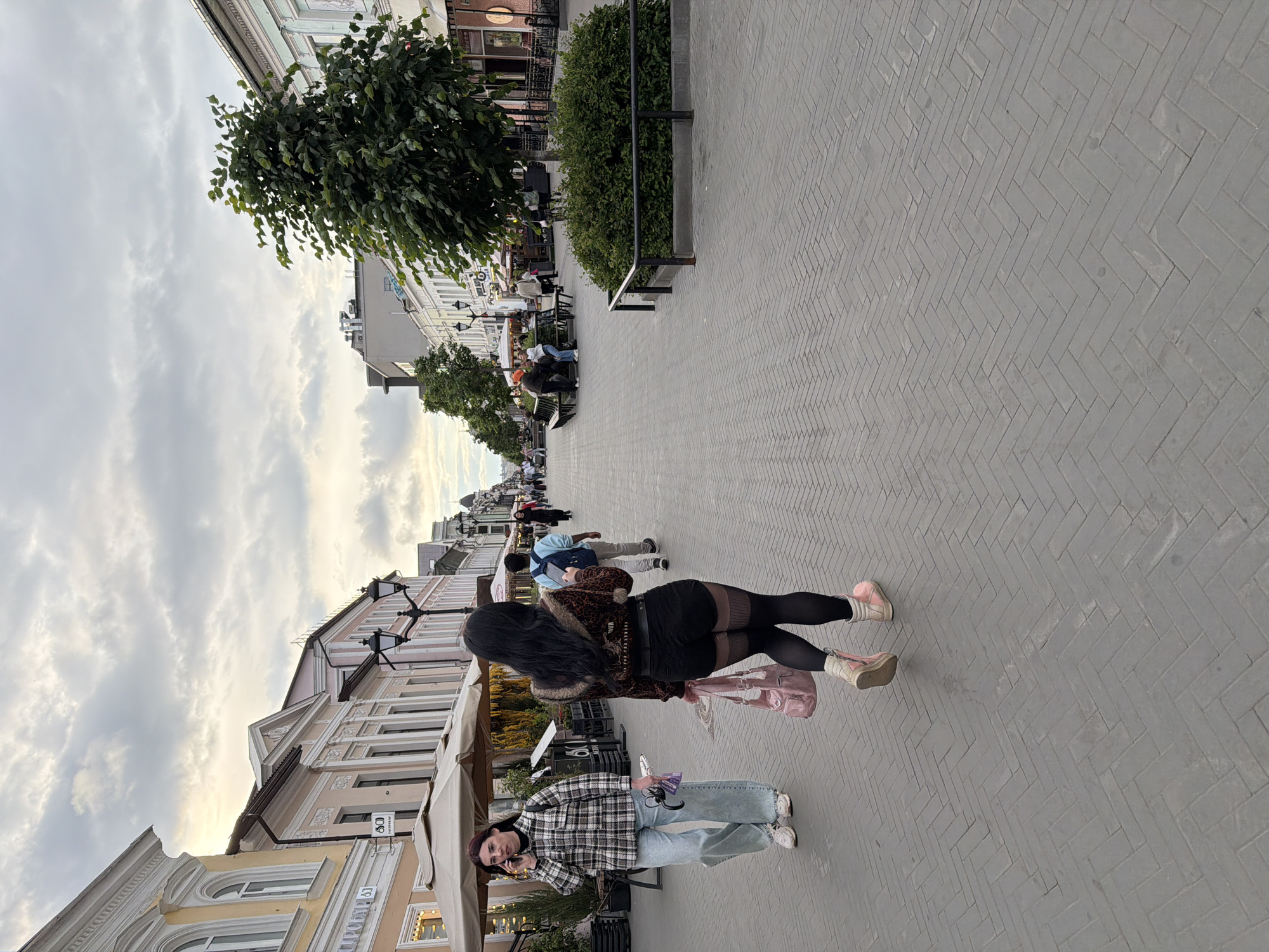 The image depicts a pedestrian zone in Kazan, Russia, identified by the architecture of buildings and the style of street furniture. The street is paved with grey herringbone-patterned blocks. Buildings line both sides of the street with shops and cafes, identifiable by awnings.
Several people are present, including two women in the foreground. One is wearing a plaid shirt and jeans, and is using a mobile phone. The other is wearing black tights, a brown jacket with fur trim, and pink boots, carrying a pink bag. Other pedestrians are walking further down the street. Trees and bushes line the street, with benches and low metal railings visible. The sky is overcast.