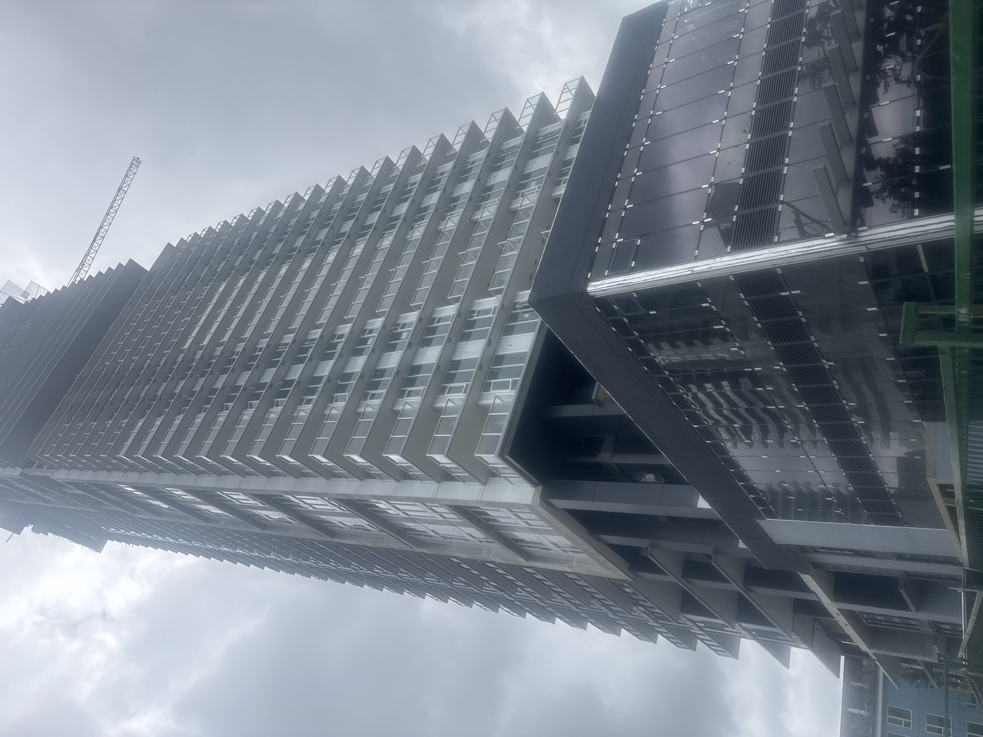 This image shows a tall, multi-story building under construction in Coquitlam, Canada. The building is characterized by its modern, angular design with numerous balconies and glass-paneled windows. A construction crane is visible extending from the upper portion of the building, indicating ongoing work. The lower section of the building features a sleek, dark-glass facade. The sky is overcast and appears to be filled with clouds. The scene is viewed from a low angle, emphasizing the height and scale of the structure. In the foreground, a green metal fence is partially visible, suggesting a construction site perimeter.