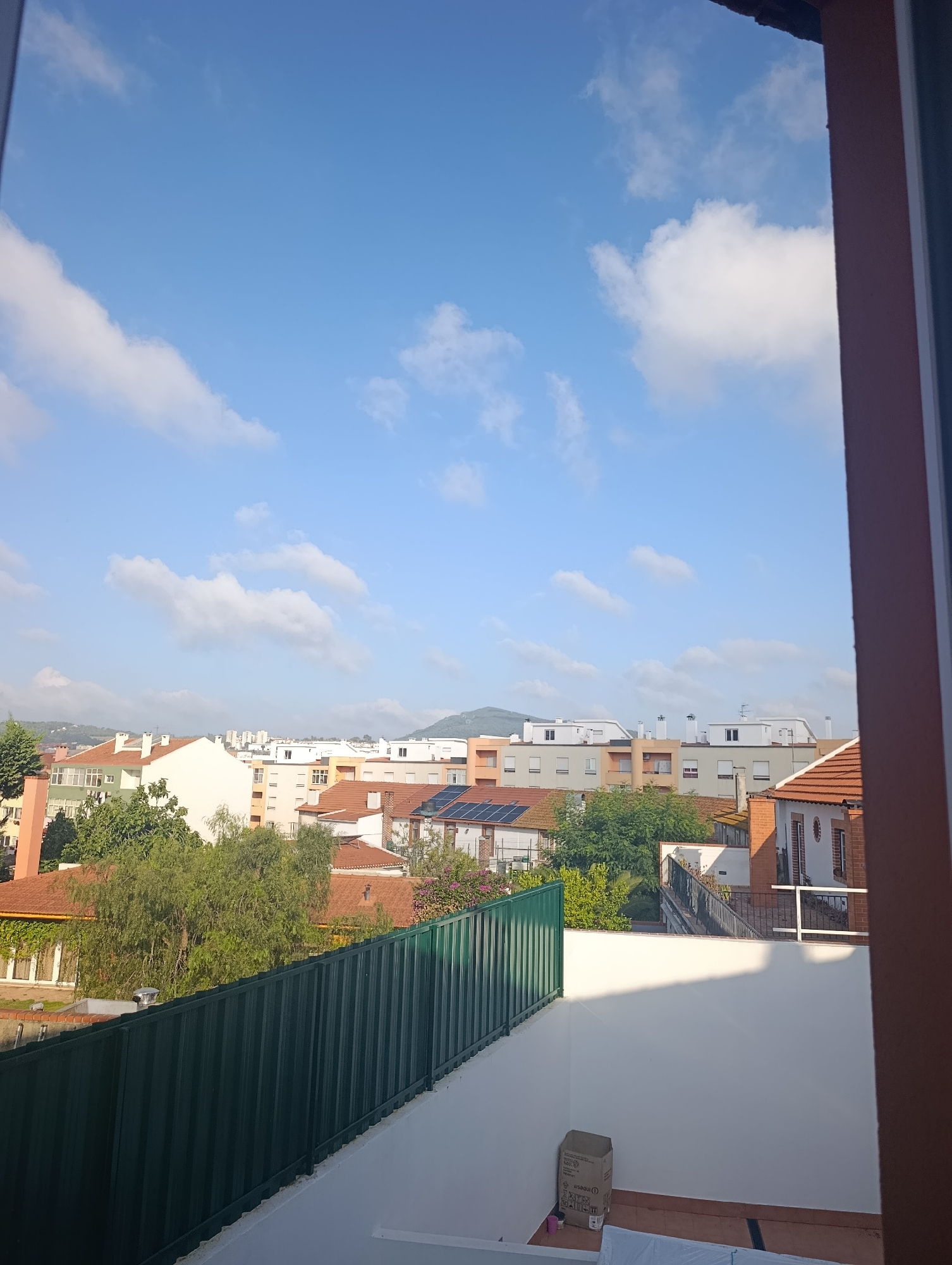 The image depicts a view from an elevated vantage point, looking out over a residential area in Setúbal, Portugal. The sky is a bright blue with scattered white clouds. In the mid-ground, a collection of multi-story buildings with red-tiled roofs and light-colored facades are visible, some with balconies and chimneys. Green trees are interspersed among the buildings. In the foreground, a dark green metal fence runs horizontally across the frame, separating a white outdoor area from the view of the city. A cardboard box and a small pink container are present on the white surface in the lower right quadrant. The overall scene suggests a residential neighborhood on a clear day.