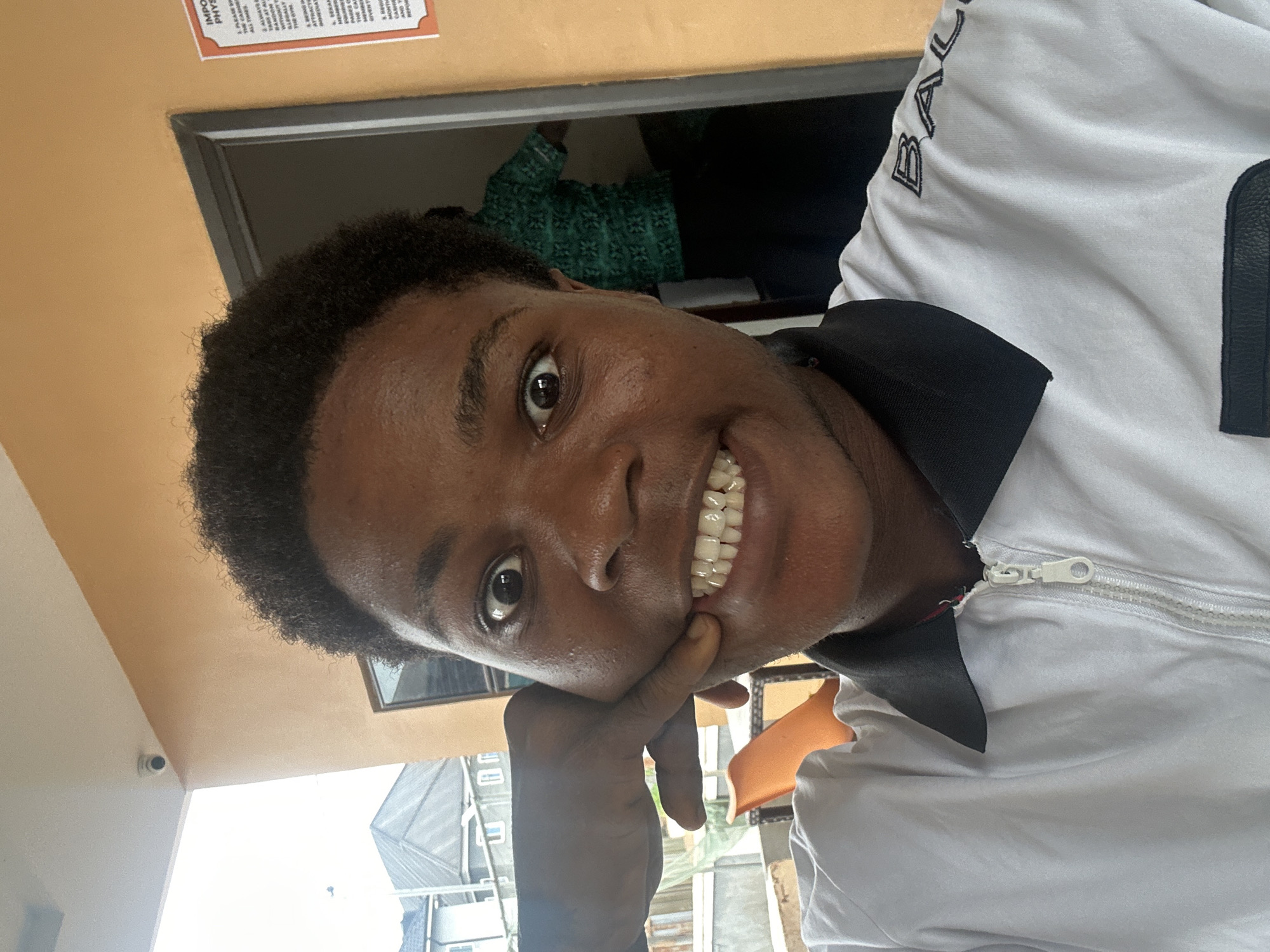 A young Black person is taking a selfie with their right hand pulling their cheek to the side and displaying their teeth. The person is wearing a white polo shirt with a black collar and a white zipper. In the background, a person in a green patterned shirt is visible through an open doorway. A sign with text is mounted on the orange wall to the right of the doorway. The person's left hand is partially visible, resting on their cheek. The scene appears to be outdoors or in a semi-open space, possibly near a building with an orange wall and a white overhang. The context suggests this could be an establishment within Ihiagwa, Nigeria, given the prompt's specific location.