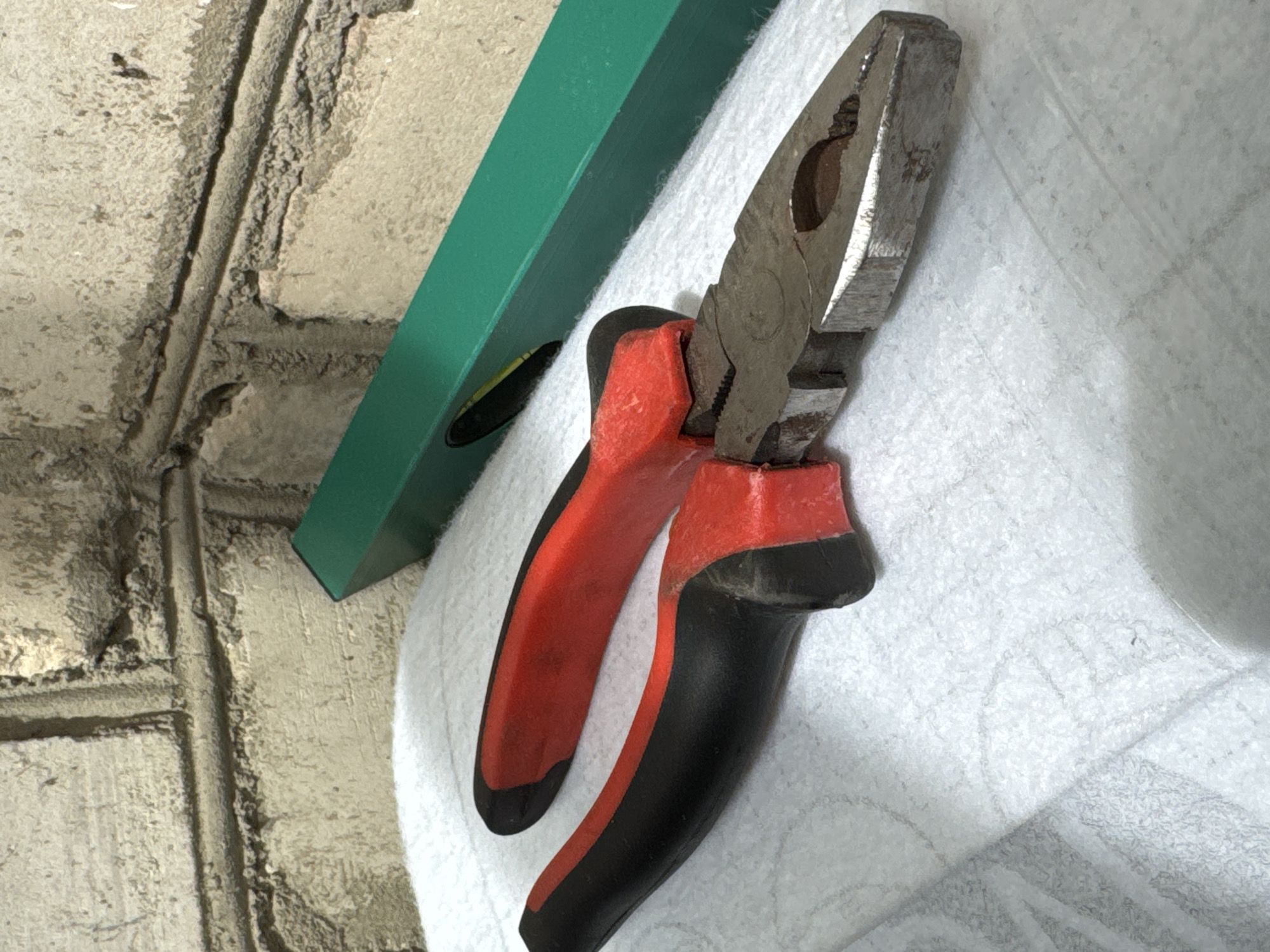 In a dimly lit, indoor environment with a textured brick wall in the background, a pair of pliers with red and black handles lies on a white surface. The pliers are angled, with the jaws slightly open, revealing their metallic components. To the right of the pliers, a green spirit level is visible, partially obscured by the pliers. The white surface appears to be a fabric or a disposable cloth, showing some wrinkles and texture. The overall scene suggests a workspace or a storage area for tools. The location in Яромінскі сельскі Савет, Belarus, is noted, but no specific urban features or context are depicted in the image itself.