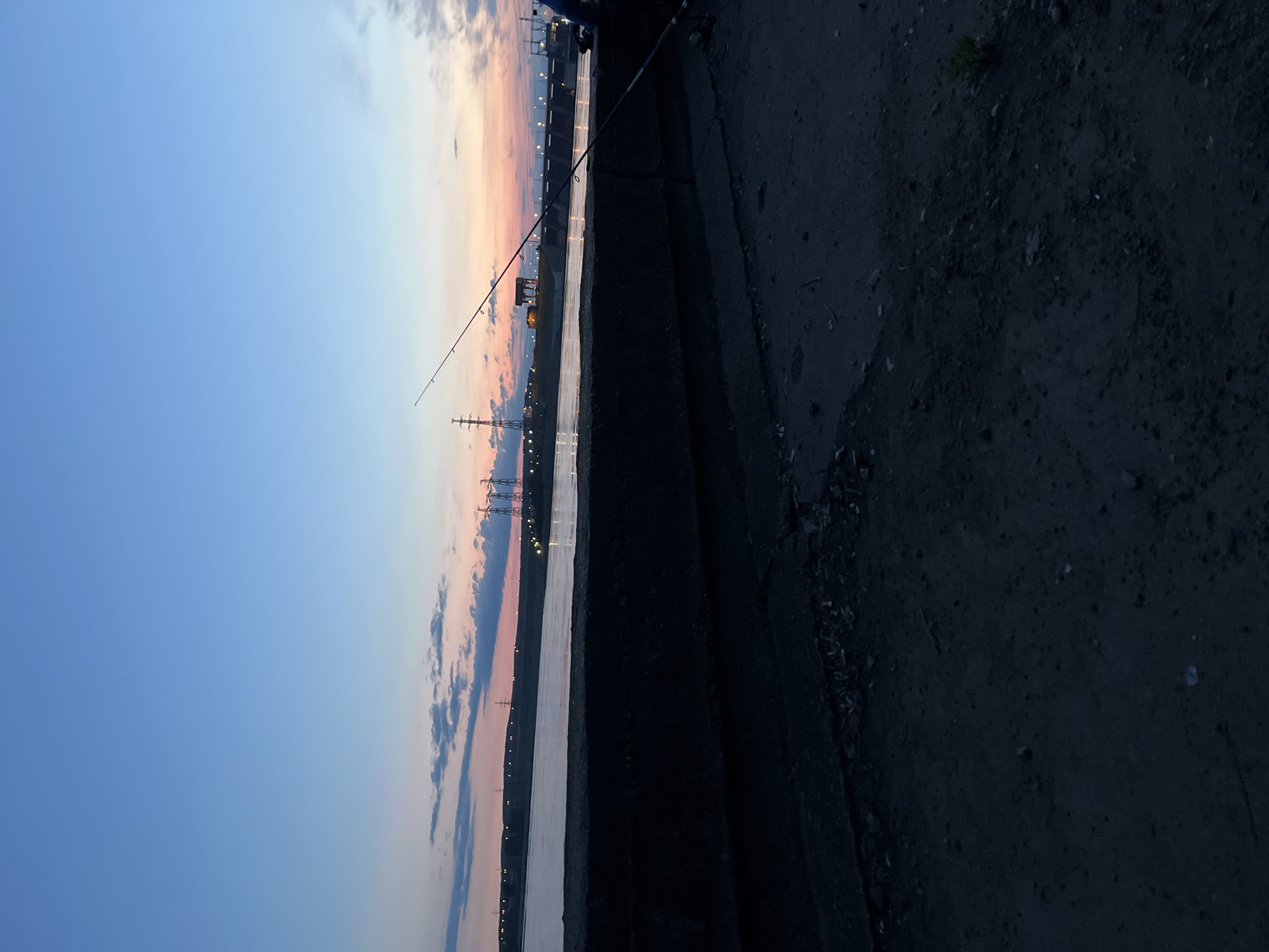 This image captures a tranquil twilight scene, likely at dusk, along a body of water in Chaikovsky, Russia. The sky dominates the upper portion of the frame, displaying a gradient from deep blue at the top to soft pink and orange hues near the horizon, accented by scattered clouds.
In the mid-ground, a calm expanse of water reflects the fading sky and distant lights. A fishing rod is prominently visible, extending from the right foreground over the water, suggesting an ongoing or recently concluded fishing activity. The foreground on the right side consists of a dark, textured embankment or shoreline, appearing to be made of concrete or packed dirt, with subtle details of small rocks and sparse vegetation.
Across the water, on the distant bank, a series of industrial structures, possibly power line towers or utility poles, are silhouetted against the colorful horizon. Several small lights are visible along the far bank, adding a subtle glow to the distance. The overall low light conditions and vibrant sky colors create a peaceful and quiet atmosphere. There is no visible text in the image.