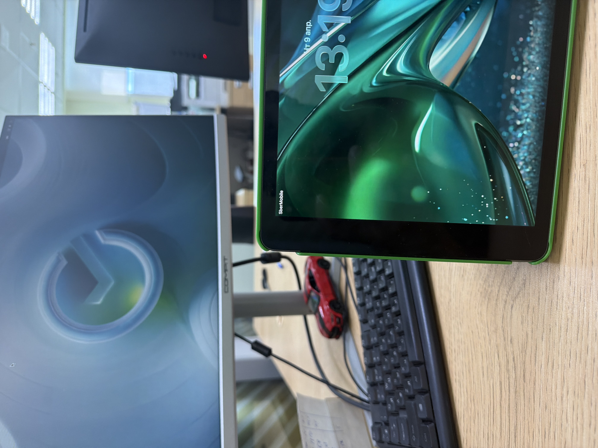 The image shows a desk with a computer monitor displaying a circular logo, a black keyboard, a red toy car, and a green tablet showing the time 13:19 and the date April 9. The tablet's brand is SberMobile. A black monitor is in the background. The environment appears to be an office or workspace. The location is unknown.