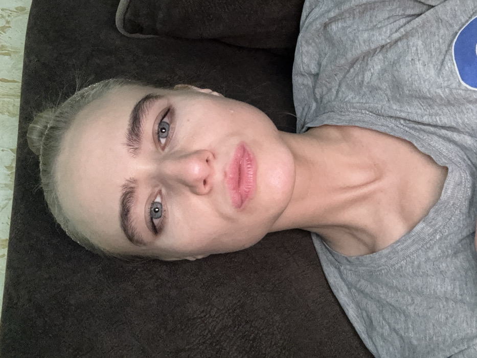 A pale-skinned person with light blue eyes and thick eyebrows is lying down, turned slightly to the side. The hair is pulled back in a bun. The person wears a gray T-shirt. The neck appears slender, with visible bone structure. The background includes a brown textured surface, possibly a couch or bed. The lighting is somewhat dim and casts subtle shadows on the face and neck.