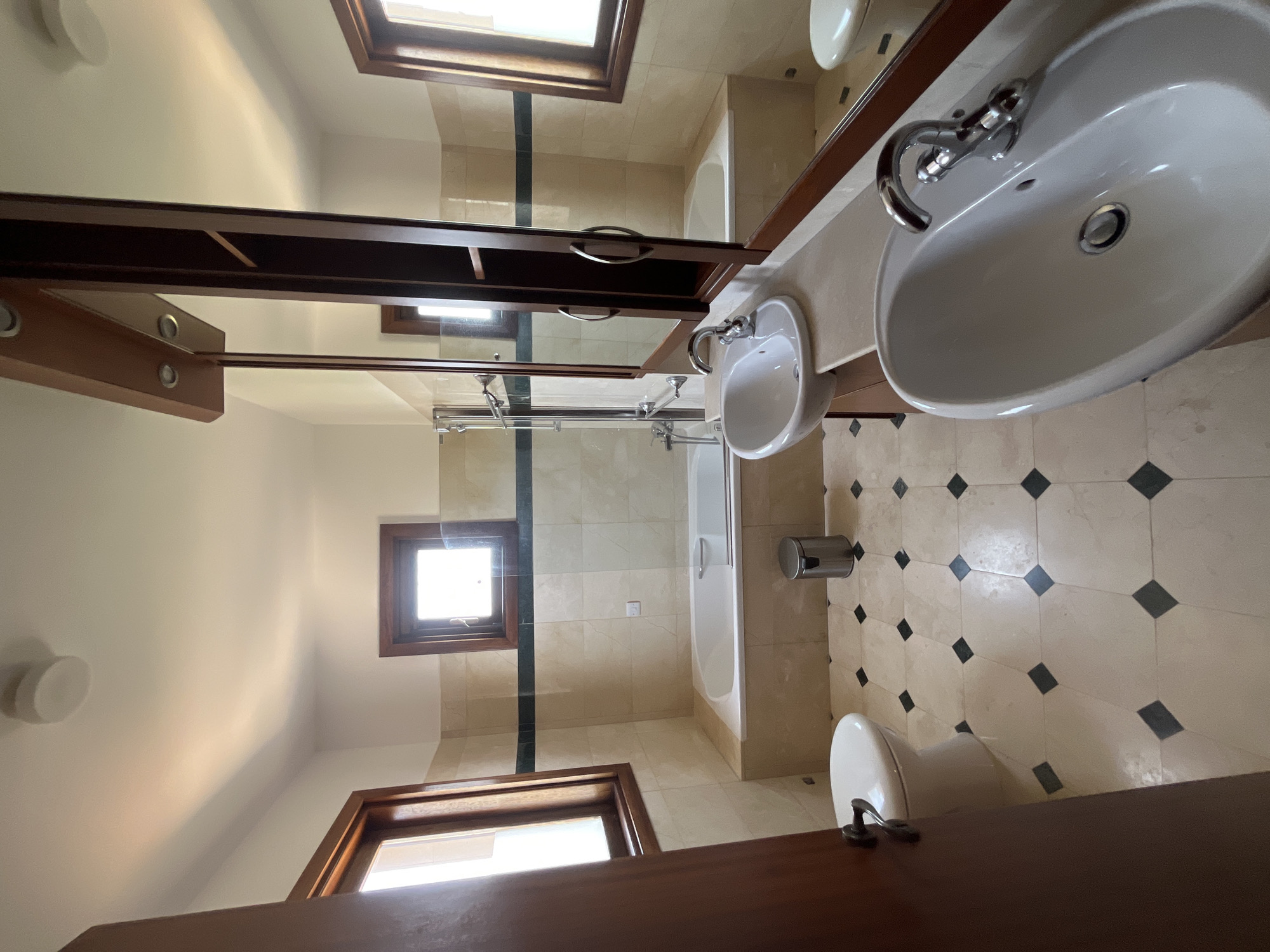 The image depicts a well-lit bathroom with beige tiled walls and floors featuring a pattern of dark diamond-shaped tiles. On the left, a white toilet with a chrome handle is visible, partially obscured by a dark brown door frame. Adjacent to the toilet is a large bathtub, also white, set against the tiled wall. A glass shower screen with a chrome frame is positioned over the tub. A stainless steel trash can stands near the bathtub.
The central and right side of the image showcases a vanity area with two white sinks. The vanity top is a light-colored countertop. Above the sinks, a wooden cabinet with mirrored doors extends upwards, featuring integrated lighting. One of the sinks has a chrome faucet. A large mirror reflects the opposite side of the bathroom, showing another window and the continuation of the tiled walls and bathtub.
Two rectangular windows with wooden frames are present in the bathroom; one on the left wall and another on the right wall, allowing natural light to illuminate the space. The overall impression is of a clean and functional bathroom, likely located within a residential property in Aphrodite Hills, Cyprus.