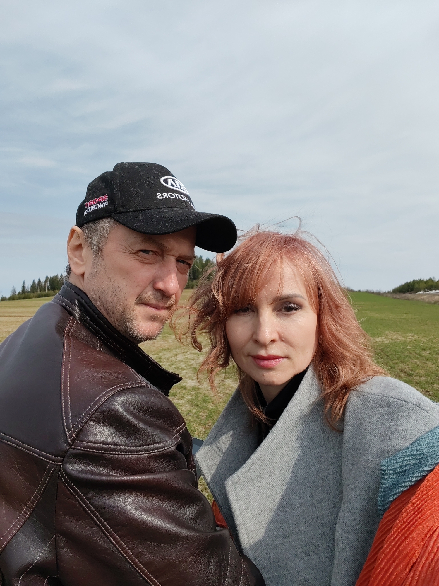 The image shows a selfie of two people in a field in what appears to be a hilly, rural area. The sky is partly cloudy. A patch of trees can be seen on a hill in the distance. The foreground consists of a grassy field.
The man on the left is wearing a black baseball cap with the word "MOTORS" emblazoned on it in reverse, along with a dark brown leather jacket. He has short, graying hair and facial hair. He is looking directly at the camera.
The woman on the right has reddish-brown, wavy hair and is wearing a gray wool coat over a black turtleneck. Part of an orange pleated garment is visible beneath the gray coat. She is also looking at the camera.
The location is Нечкино, Russia.