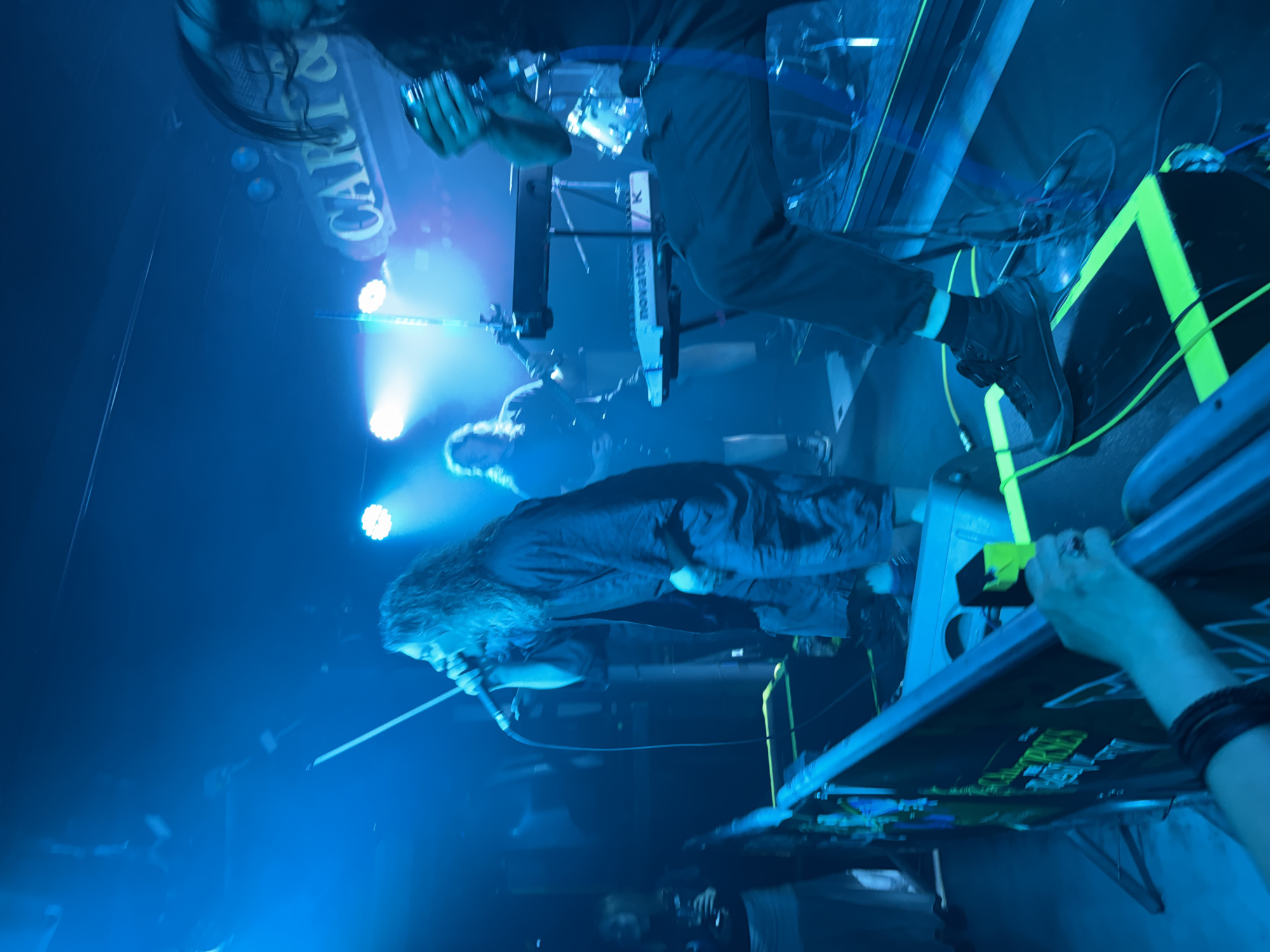The image shows a music performance venue in Unknown, Unknown. The scene is lit with blue stage lights. A singer with long hair is leaning into a microphone. Behind him, another musician is playing a keyboard labeled "Novation." Stage equipment, including amplifiers and cables, is visible. A hand holding a camera or phone is in the foreground. The name "CARAVIA" is partially visible in the background.