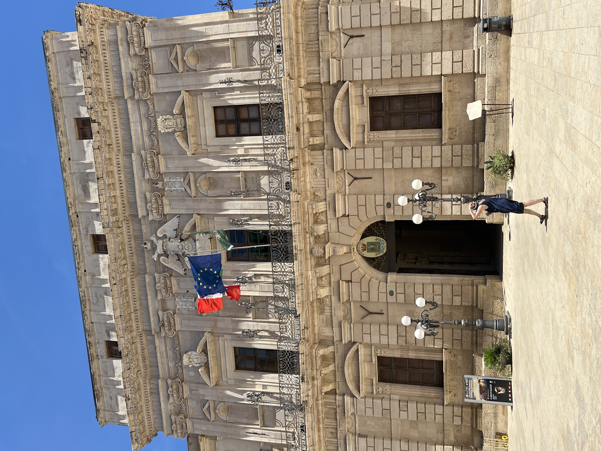 In Syracuse, Italy, the facade of a building with ornate stonework and decorative elements is visible. The building features multiple windows, a balcony with an intricate iron railing, and a large arched entryway. Above the entryway, the Italian flag and the European Union flag are displayed. A single person in a dark dress is walking away from the camera in the foreground, looking up and holding a phone to their ear. Two ornate lampposts stand on the paved plaza in front of the building, along with a sign advertising an exhibition. The sky is a clear blue.