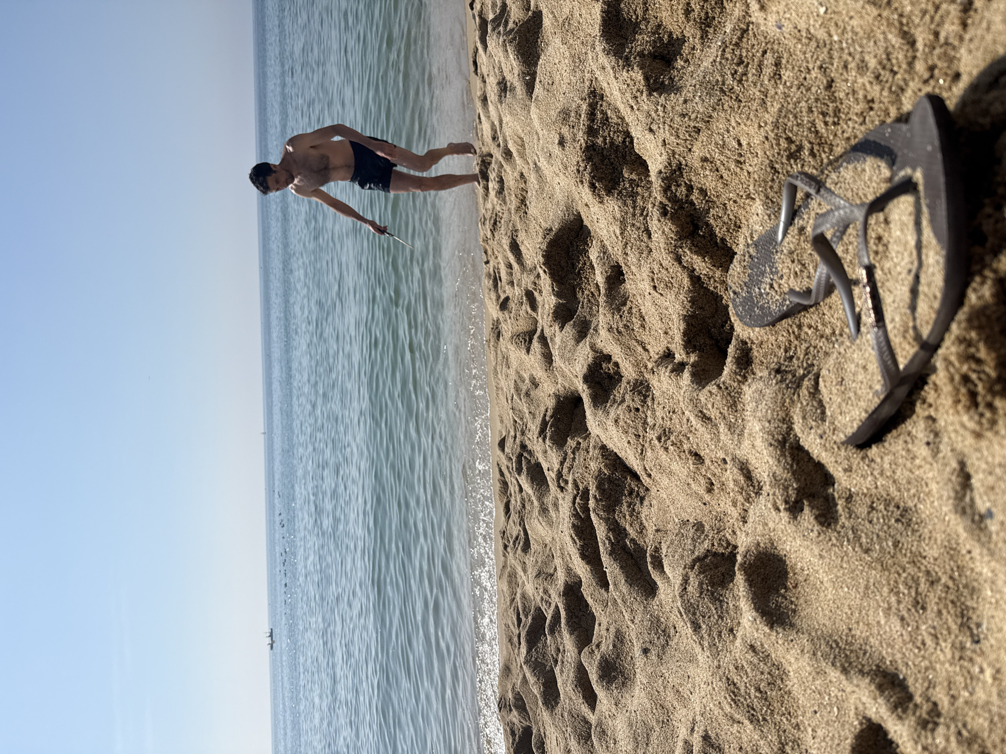 The image shows a person on a sandy beach next to the ocean. The person, a male, is wearing dark swim trunks and is bent over as if looking for something in the sand. He is holding a small, flat object in his outstretched hand. In the foreground, on the sand, is a pair of dark flip-flops. The ocean is calm with gentle waves lapping at the shore. The sky is clear and blue, suggesting it is daytime and fair weather. The scene is likely taking place in a sunny, warm environment, consistent with a beach location in Barcelona, Spain. The image is oriented vertically, with the horizon line running across the middle of the frame. There are no visible text elements in the image.