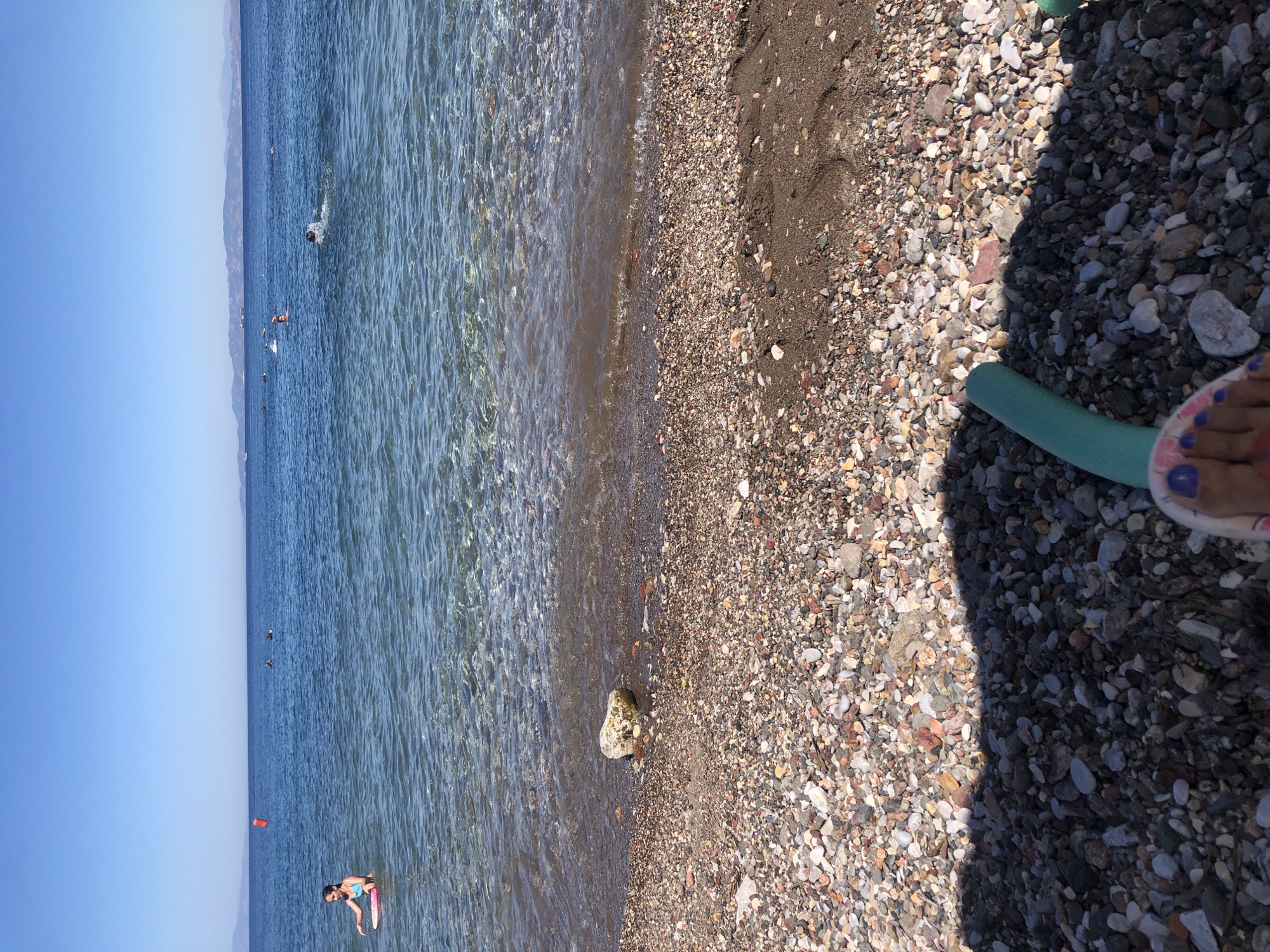 The image shows a view of a rocky beach and the sea. In the foreground, a person's feet are visible, along with a green pool noodle partially shaded by an unseen object. The beach is covered in small, multi-colored pebbles. The sea is calm and clear, with gentle waves lapping at the shore. Several people are visible swimming and playing in the water further out. In the distance, a hazy coastline with mountains can be seen under a clear blue sky. The image appears to be taken from a low vantage point on the beach. The location is identified as Karaburun, Turkey, suggesting a coastal setting in this region.