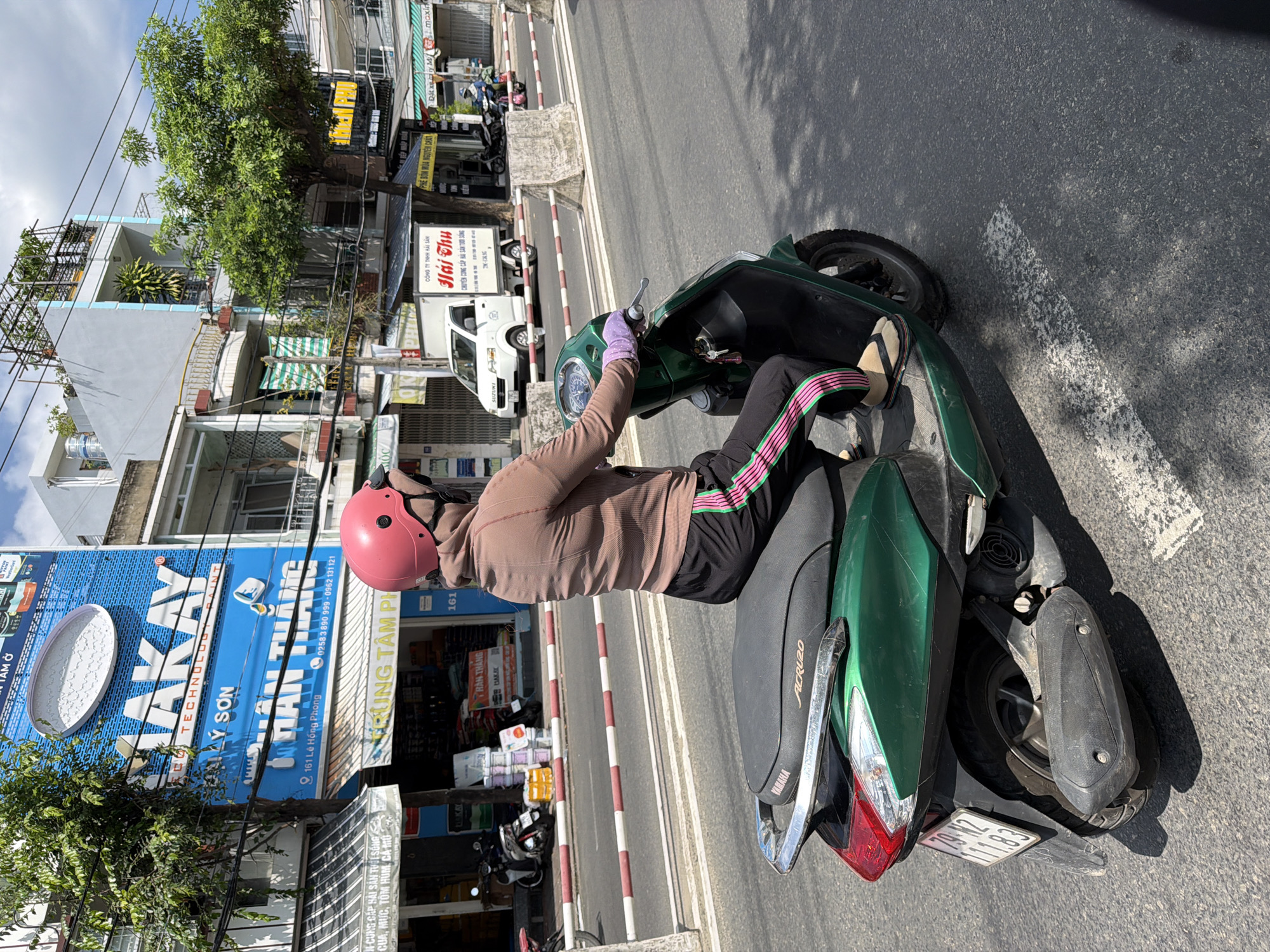 A woman wearing a pink helmet and a brown long-sleeved shirt rides a green Yamaha scooter on a city street in Nha Trang, Vietnam. She wears black pants with pink and green stripes and tan flip-flops. The scooter is in motion, with the rear wheel visible, and has a license plate reading "79-N2 111.83". In the background, buildings line the street, including one with a large blue sign that says "MAKAY TECHNOLOGY PAINT". A white Suzuki truck and other parked motorcycles are also visible. Red and white striped barriers are on the side of the road. The sky is partly cloudy. The image appears to have been taken from another vehicle traveling on the same road.