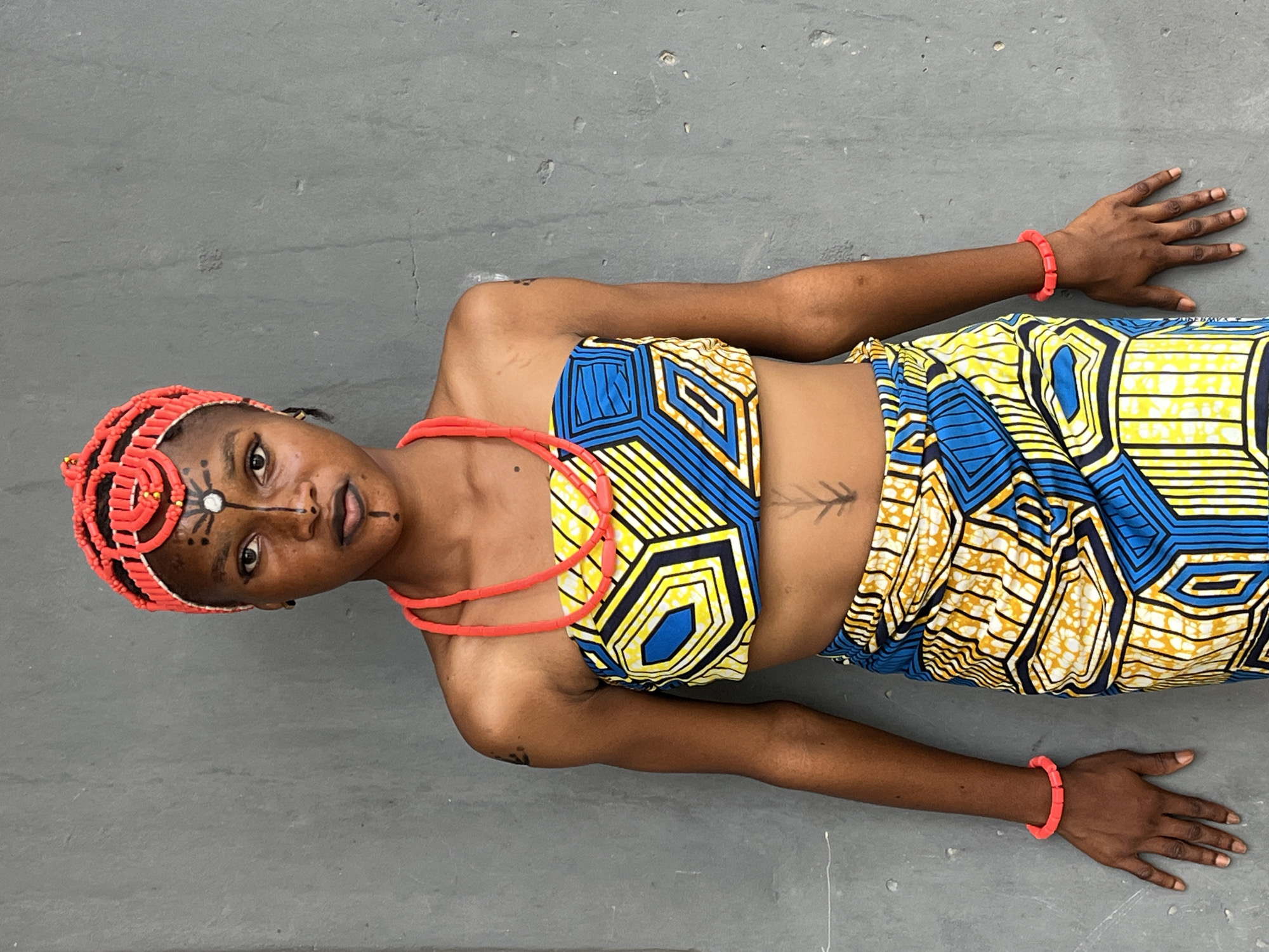 A person lies on a gray surface. The person, likely female, wears a colorful patterned two-piece outfit in blue, yellow, and white, along with red beaded headwear, necklace, and bracelets. Facial markings are visible. The background is a plain gray surface, potentially concrete. No specific details indicating Badagry, Nigeria are readily discernible.