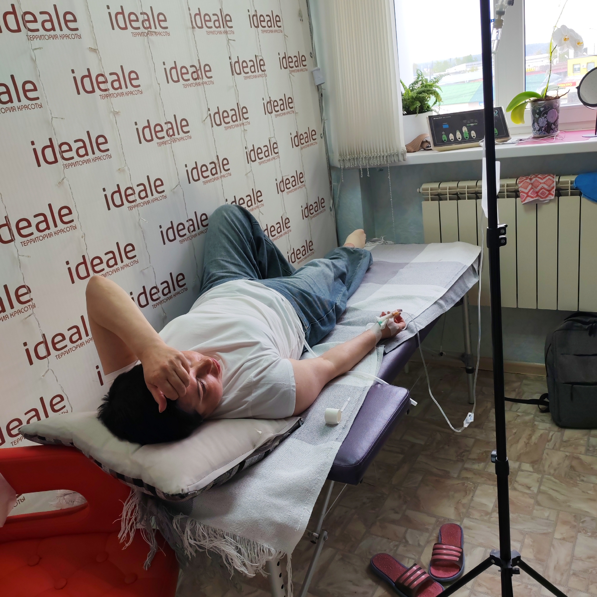 A person lies on a treatment table in a room of a beauty salon or medical clinic, evidenced by the branding "ideale ТЕРРИТОРИЯ КРАСОТЫ" (ideale BEAUTY TERRITORY) repeatedly displayed on the wall. The person is receiving an intravenous drip in their arm. Medical equipment and a black tripod are visible near the treatment table. Red and pink slides are on the floor near the base of the tripod. A backpack rests near the treatment table. A window with plants sits along the wall behind the treatment table. Based on the OCR, this picture might be from Chaikovsky, Russia.
