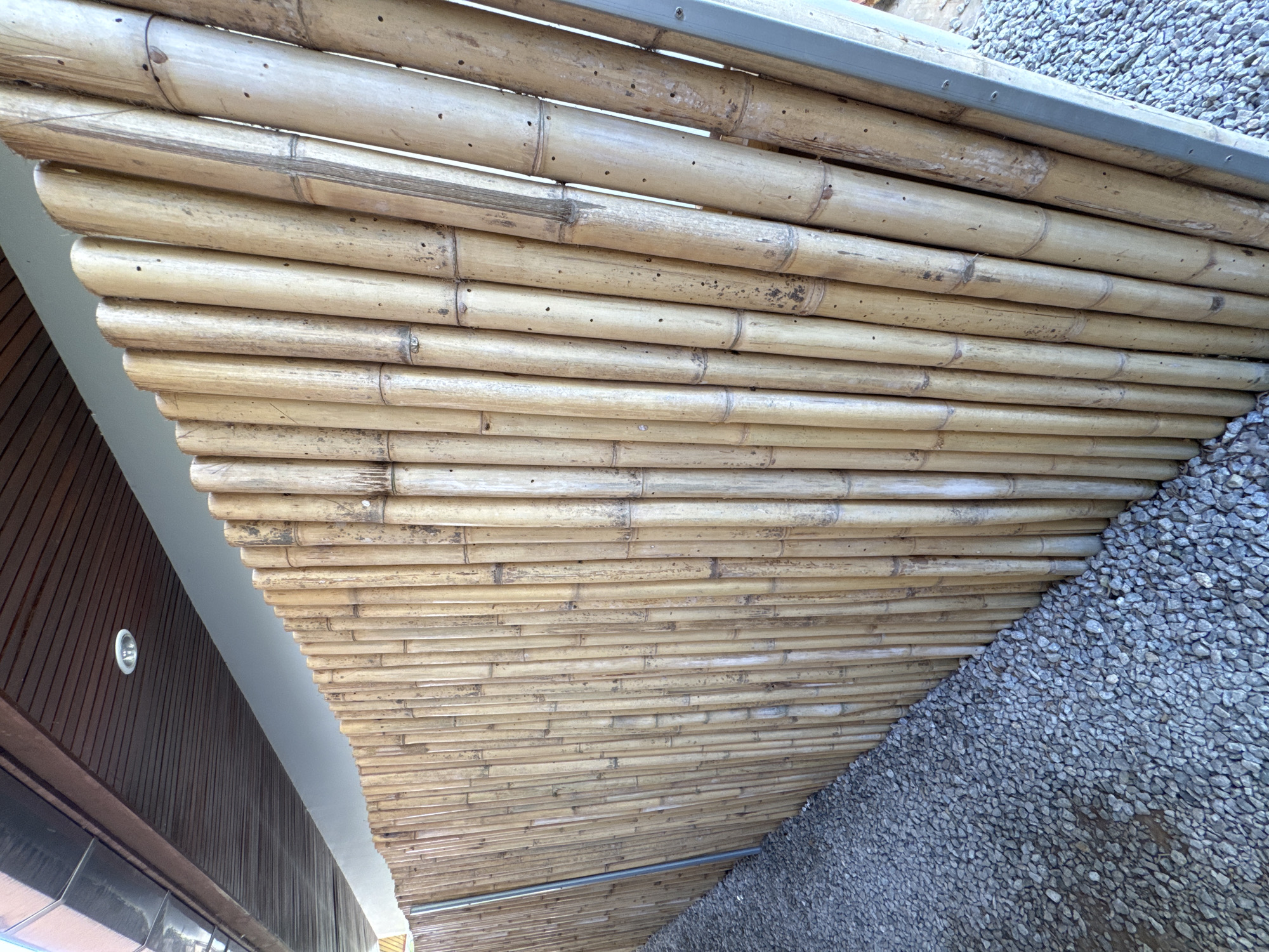 Here's a detailed description of the image:
The image shows a close-up view of a bamboo ceiling or overhang, possibly part of a building structure. The bamboo is arranged horizontally, forming a textured surface. The color of the bamboo is light brown/tan.
To the right of the bamboo, there is a section of gravel or small stones, indicating an exterior environment. To the left, there is a building with wooden paneling and a light fixture.
The presence of bamboo as a building material and the gravel pathway suggest an outdoor or semi-outdoor space. Based on the location provided (Rawai, Thailand), the style of the building may reflect Southeast Asian architectural influences.