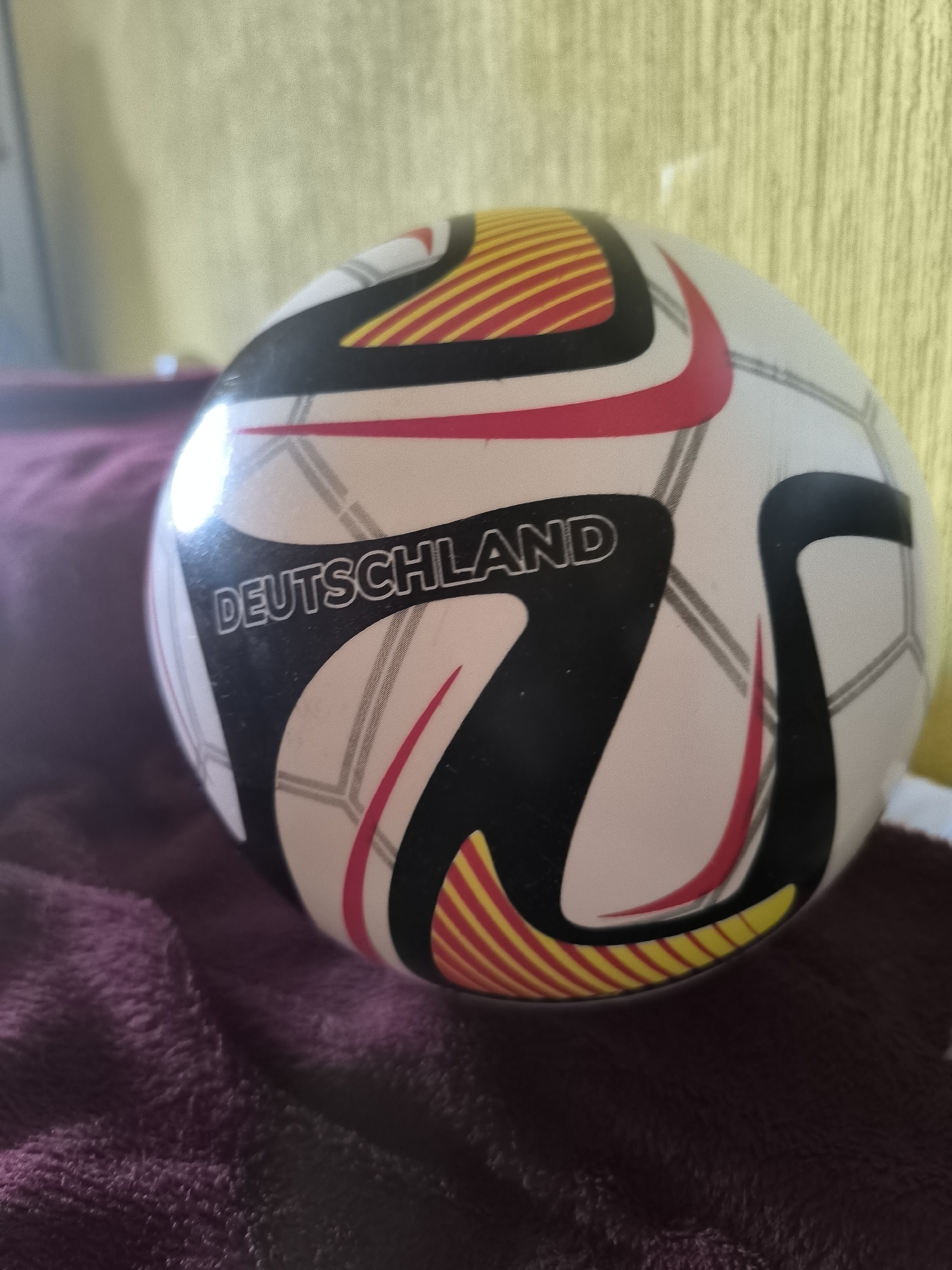 This is a close-up, eye-level, medium shot of a soccer ball. The ball is white with black, red, and yellow designs. The word "DEUTSCHLAND" is visible in large, white letters on a black panel. The ball is resting on a dark purple, plush surface, possibly a blanket or piece of furniture. In the background, a light yellow wall with a subtle vertical texture is visible. The lighting appears to be artificial and casts a bright reflection on the top left of the ball. No people are visible in the image, and no specific activity or event is depicted beyond the static presentation of the object. The setting is indoors and the time of day is indeterminate.