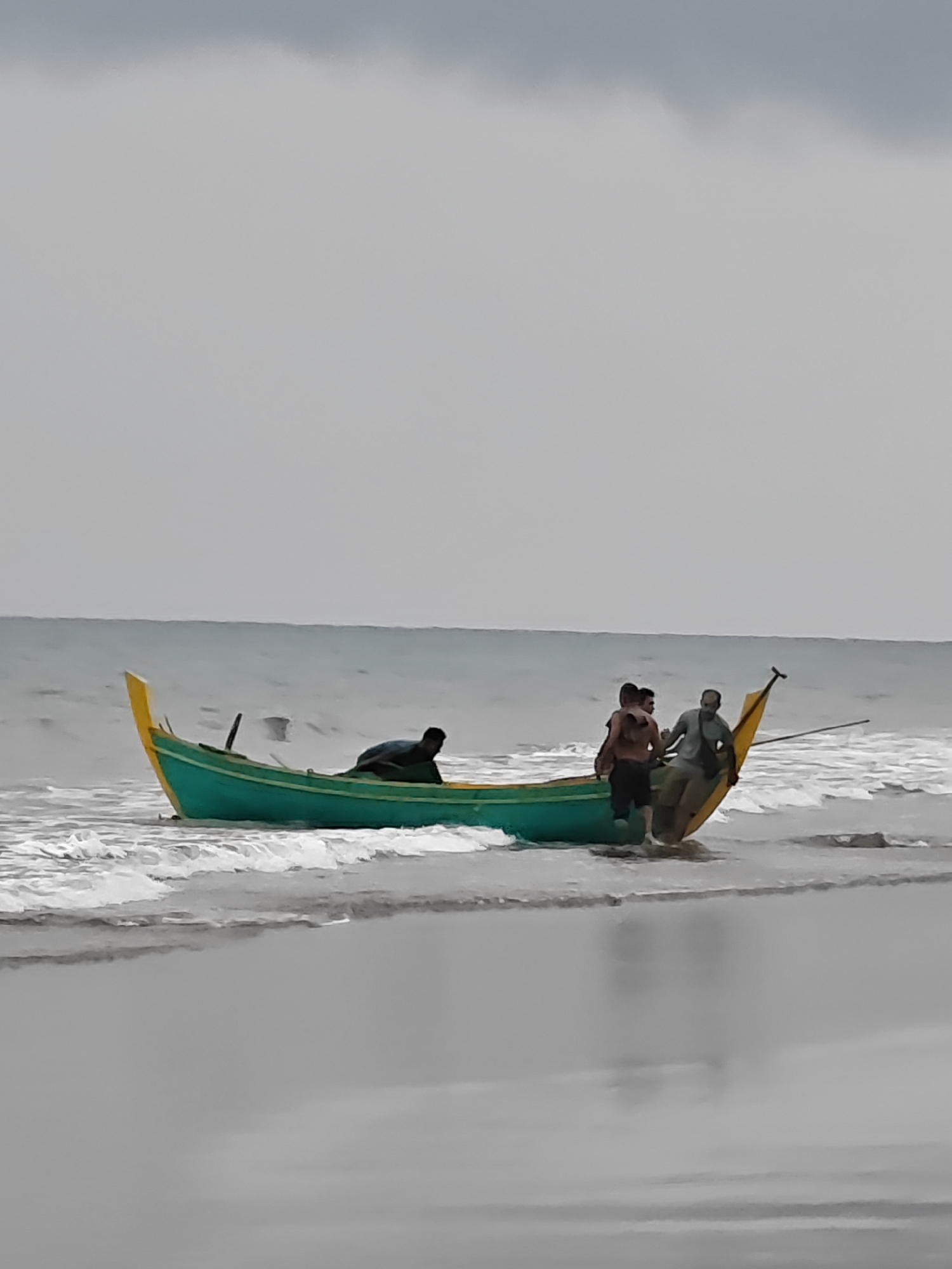 A green and yellow fishing boat is partially beached on a shoreline with waves lapping at its hull. Three men are visible in and around the boat. One man, wearing a dark shirt, is seated in the rear of the boat. Two other men are standing in the boat, one in the middle and another towards the front. The sky is overcast and the sea appears choppy. This scene is located in Curehtunong, Indonesia.
