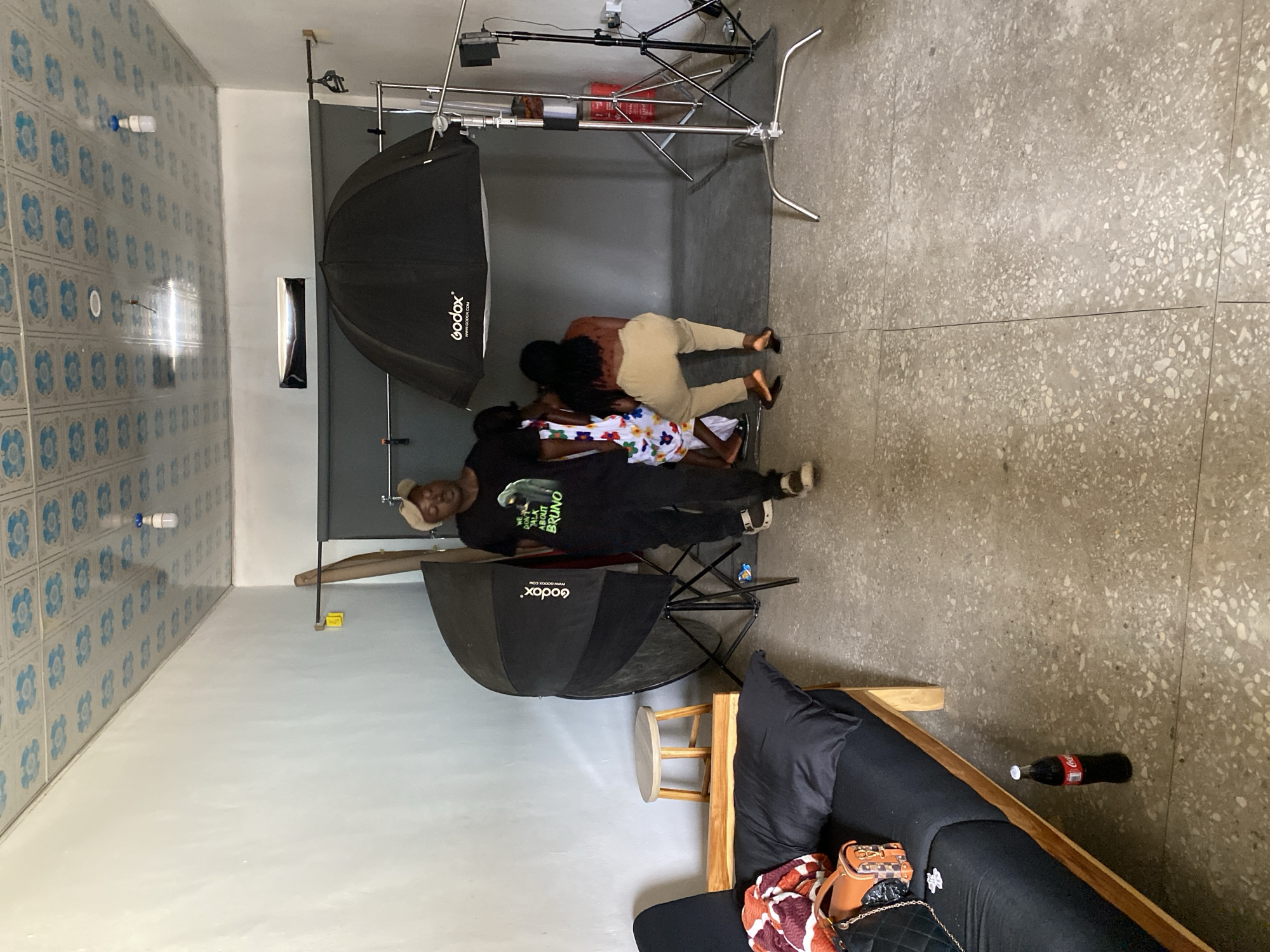 The image depicts a scene in what appears to be a photography studio in Gura Top, Nigeria. There are two people lying on the floor in front of a gray backdrop and beneath two large softbox lighting setups with Godox branding. One individual is wearing tan pants and has long, dark hair. The second is wearing a white dress with a floral pattern. A man wearing a black t-shirt, black pants, and a light-colored hat is standing near one of the softboxes. A lighting stand is set up behind the backdrop. Other objects in the room include a small wooden stool, a dark-colored couch with various bags and a Coca-Cola bottle nearby, and walls decorated with blue and white tiles. The floor is a speckled gray material. The room is illuminated by both the lighting equipment and overhead lighting fixtures.