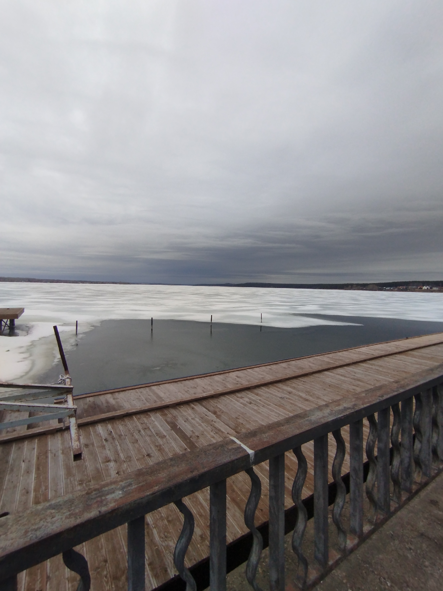 The image depicts a view of a partly frozen body of water, likely a section of the Votkinsk Reservoir, taken from a wooden pier or bridge in Votkinsk, Russia. The sky is overcast with a gray cloud cover. In the distance, the far shoreline is visible with a few buildings and treelines. The water closer to the pier is only partially frozen, with sections of open water visible amidst the ice. The foreground features the wooden planks of the pier and a metal railing. No people are visible in the frame.
