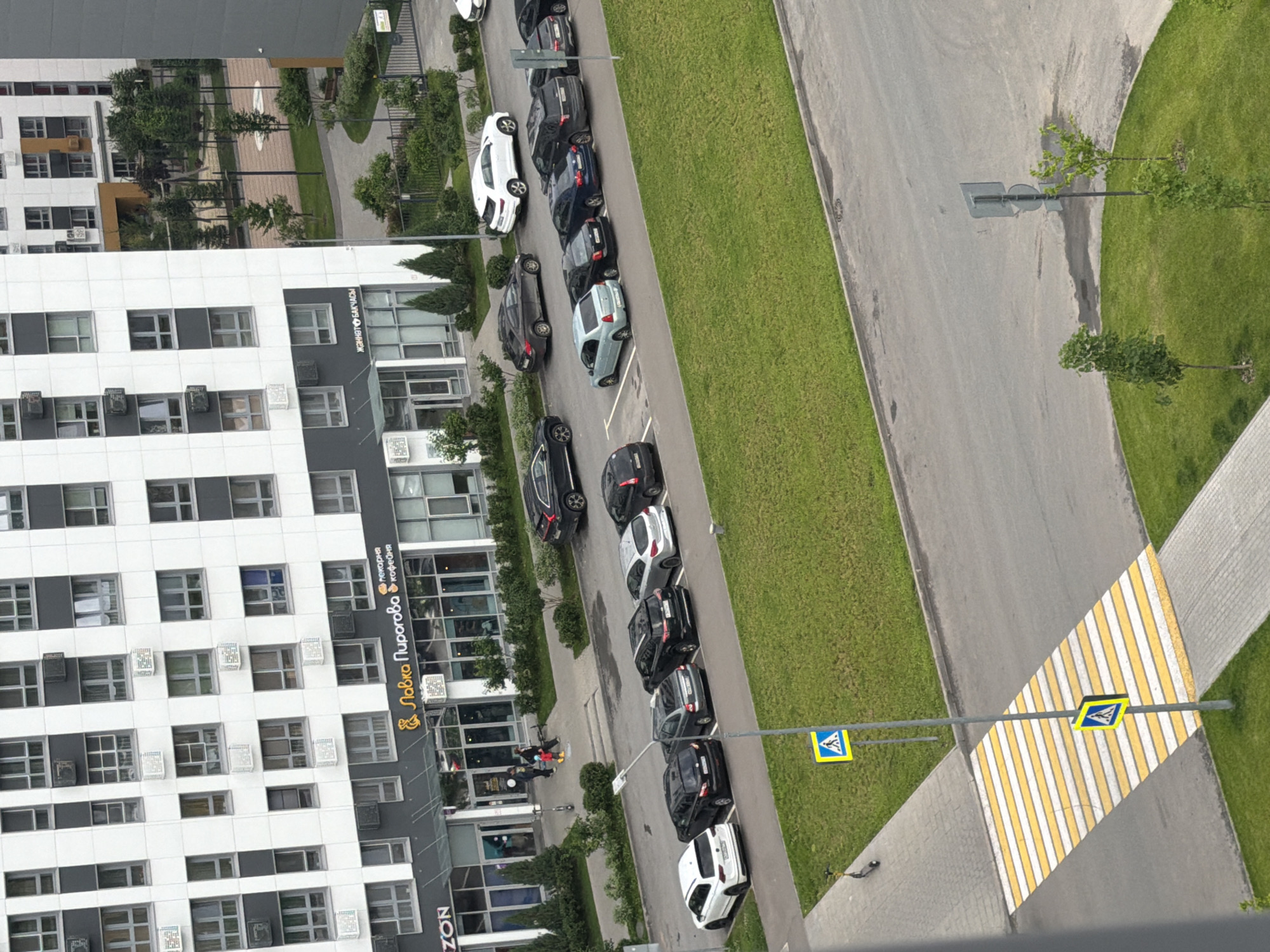 An aerial, eye-level shot shows a modern apartment building with a gray and white facade, featuring numerous windows and air conditioning units. A commercial storefront with large windows and signage for "ZON" and "Лавка Пирогова" is visible at the ground level. Several parked cars, including white, black, and silver sedans and SUVs, are lined up in a parking lot adjacent to the building. A pedestrian crossing with yellow and white stripes is present, marked by two blue and yellow pedestrian crossing signs. A few trees and green grassy areas are also visible in the scene, along with a playground in the background. A dark-colored electric scooter is parked near the sidewalk. The scene appears to be in a residential urban area.