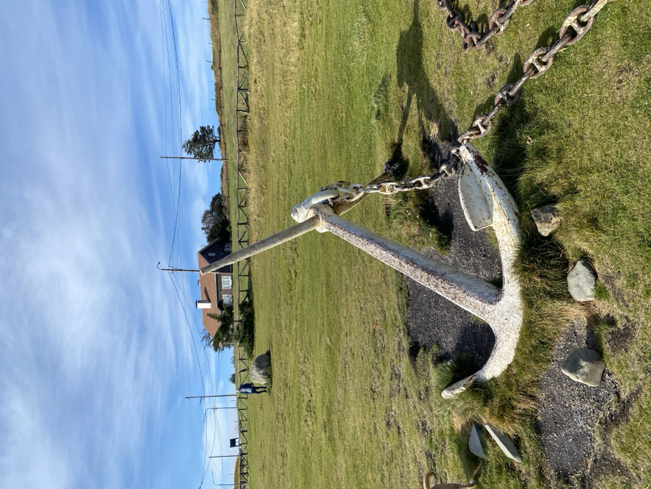 An outdoor scene in the city of Ushuaia, Argentina, is depicted under a bright blue sky with scattered cirrus clouds. A large, weathered white anchor, exhibiting rust and dirt stains, occupies the lower-center and right foreground, its fluke partially resting on dark soil and sparse green grass. A heavy, rusted metal chain extends from the anchor's stock, trailing rightward out of the frame across a patchy green lawn interspersed with small gray rocks. To the immediate left, the head and upper torso of a light-colored dog are partially visible on the grass. Further back, a person wearing a blue jacket and dark pants walks rightward across a gently sloping green field, positioned near a large, light-colored boulder. A rustic wooden fence runs horizontally across the mid-ground. In the background, an elevated terrain features a brown-roofed, dark-sided house with white window frames, accompanied by utility poles with overhead power lines and scattered trees, including one prominent tree to the right of the house. Long shadows are cast across the grass.