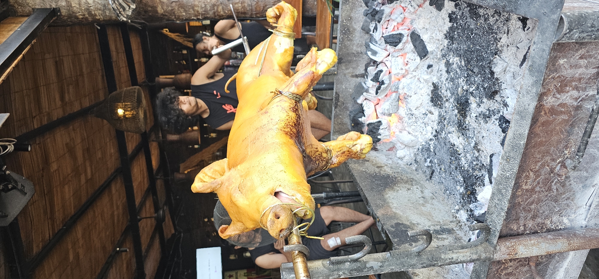 In Denpasar, Indonesia, a whole roasted pig is the focus of this angled shot. The pig, golden brown and secured with metal wire, hangs over a grill where hot coals burn. Three men attend to the pig. One man with curly hair is visible in the background, along with another man who is partially obscured, with a third tending to the pig at the grill. The scene is set in a structure with a bamboo-like wall. A light fixture hangs in the background.